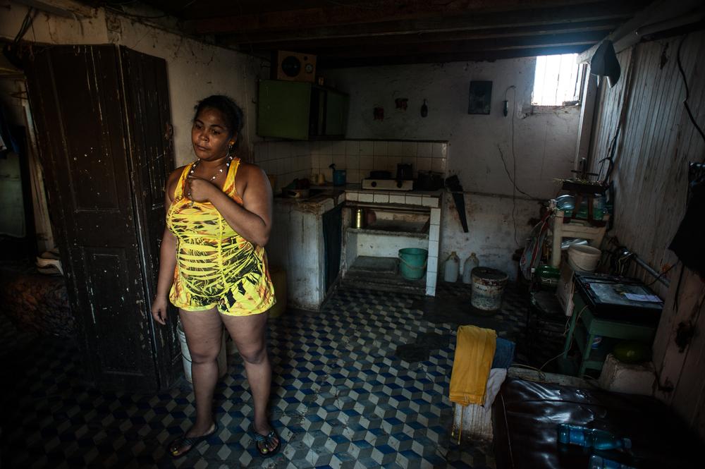 <p>A young woman stands in her kitchen. The floor of the landing in front of this apartment has caved in and large marble slabs have fallen down to the hallway below. The couple that live in this apartment have no running water.</p>