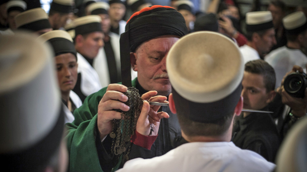 Sheikh Adrihusejn Shehu inserts a zarf into the cheek of a member of the sect [Ferdi Limani/Al Jazeera]
