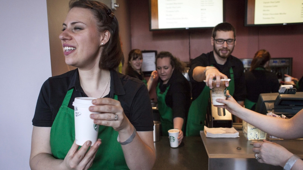 Barista in green apron holding coffee and smiling while behind her, other baristas hand coffees over the counter to customers