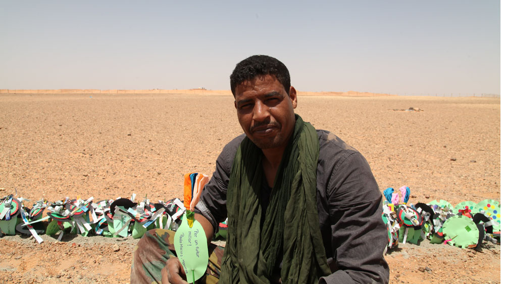 Sahrawi artist Mohamed Yeslem poses with paper peace flowers at the Moroccan-built wall [Hannah McNeish/Al Jazeera]