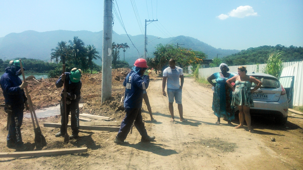 Construction work has broken the community's water piping. Residents Raquel and Helouise watch as construction workers attempt to repair the damage [Maya Thomas-Davis/Al Jazeera]