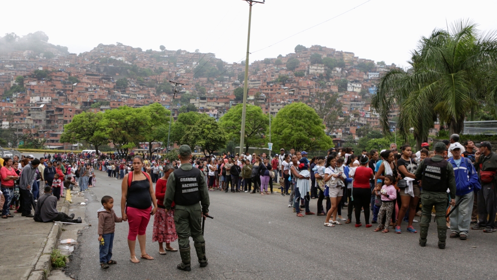 Venezuelan soldiers stand guard in a street outside a supermarket while people queue to try to buy basic food items in Caracas