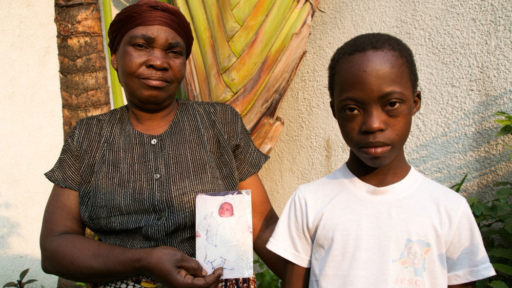 Marie Ampioje, left, stands with her son Djames Elonza, holding a photo from shortly after his diagnosis with Down syndrome [ Kait Bolongaro/ Al Jazeera]
