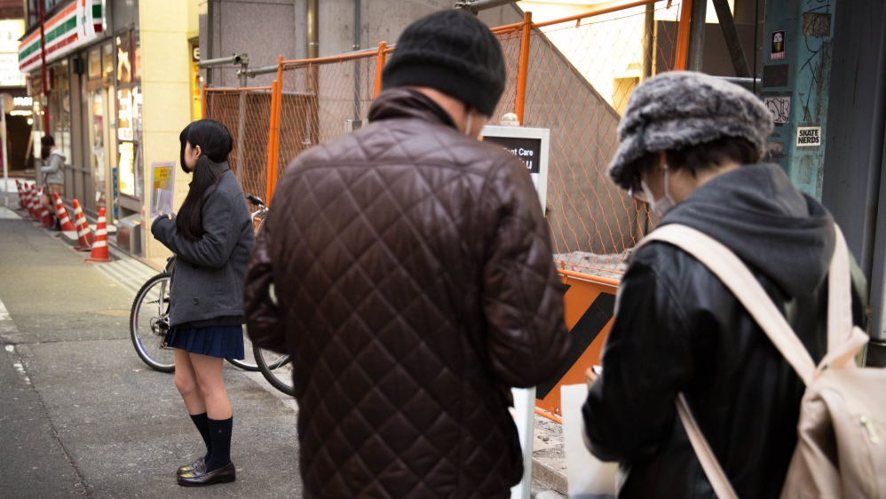 
A girl in school uniform stands on the street to gather customers for a JK cafe, while two men look at the price and service list [Shiori Ito/Al Jazeera]
