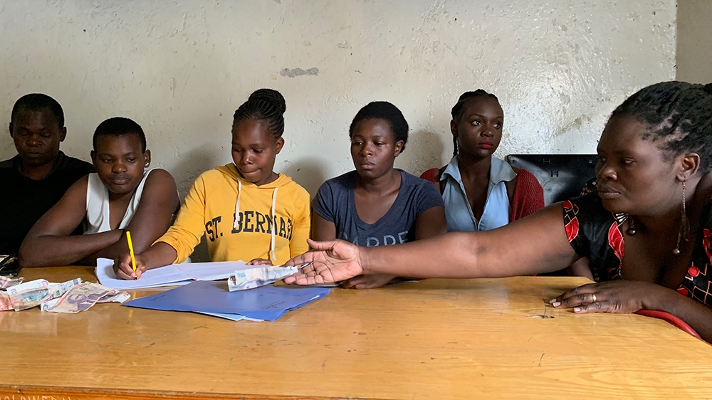Irene Obahiambo contributes her money during the 'merry-go-round'. Chair Miriam Okungu, wearing a yellow jumper third left, records her contribution [William Worley/Al Jazeera]