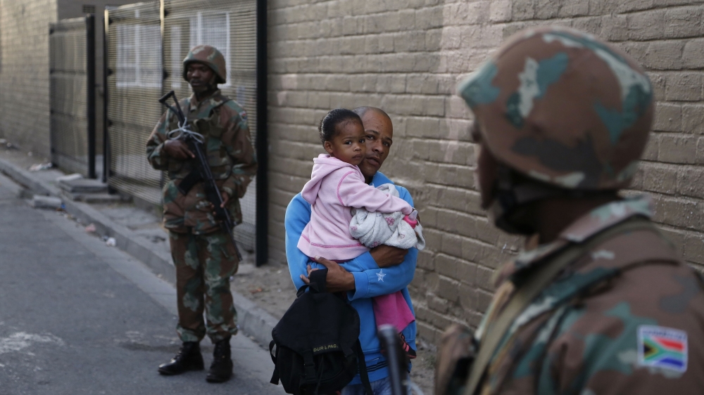 A man holding a girl walks by two South African soldiers providing security for police officers during a raid in Manenberg, South Africa Thursday, May 21, 2015