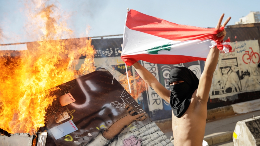 A masked protester holding a Lebanese flag walks past a burning barricade during ongoing anti-government protests in Beirut