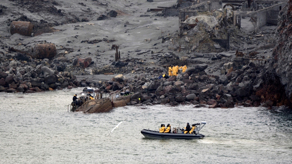 Rescuers on the White Island volcano on a mission to retrieve victims of the eruption. The land is grey and dark. There is a second group of rescuers in a dinghy on the water.
