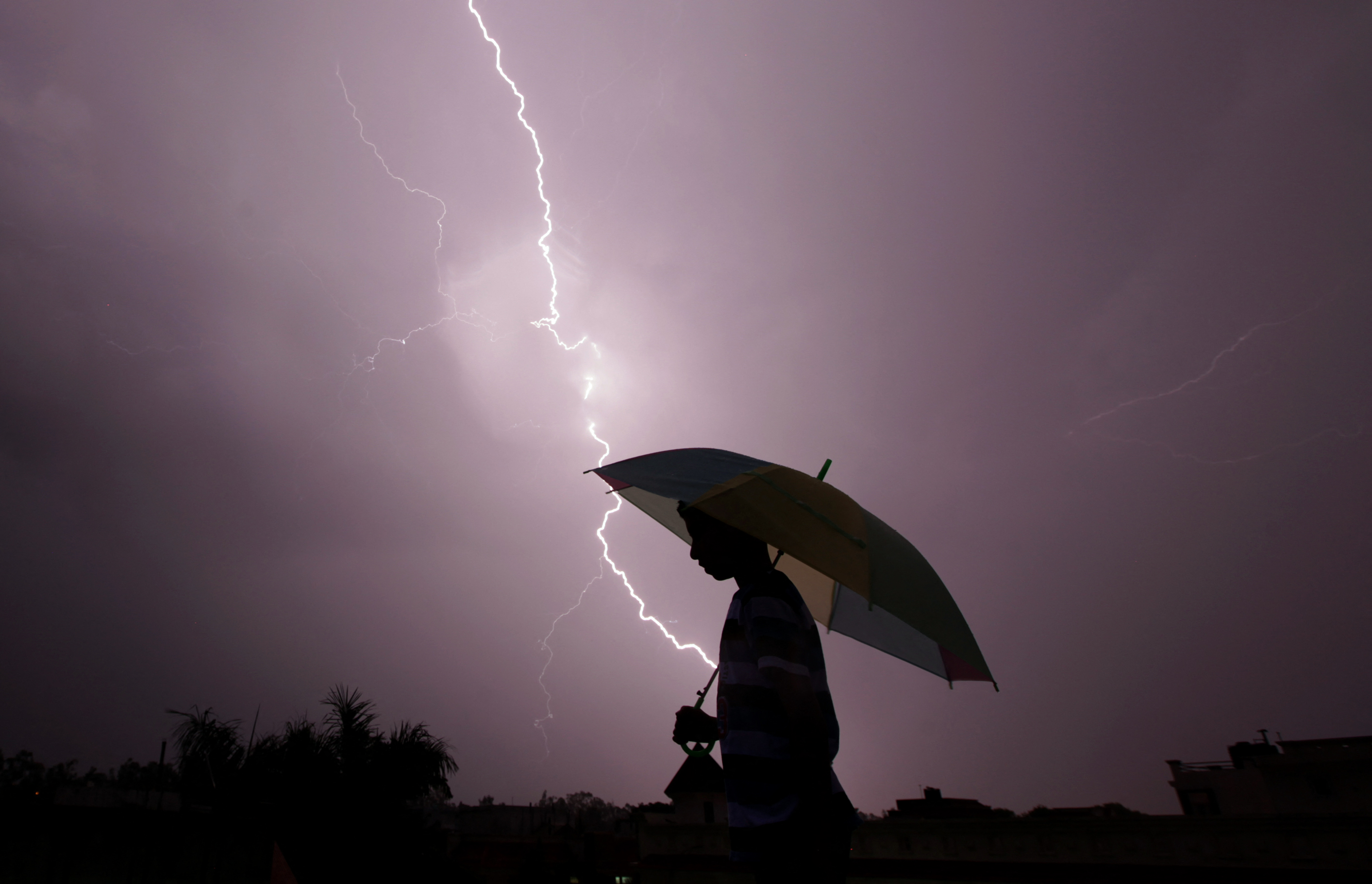 A pedestrian walks with an umbrella as lightning strikes during an evening thunderstorm