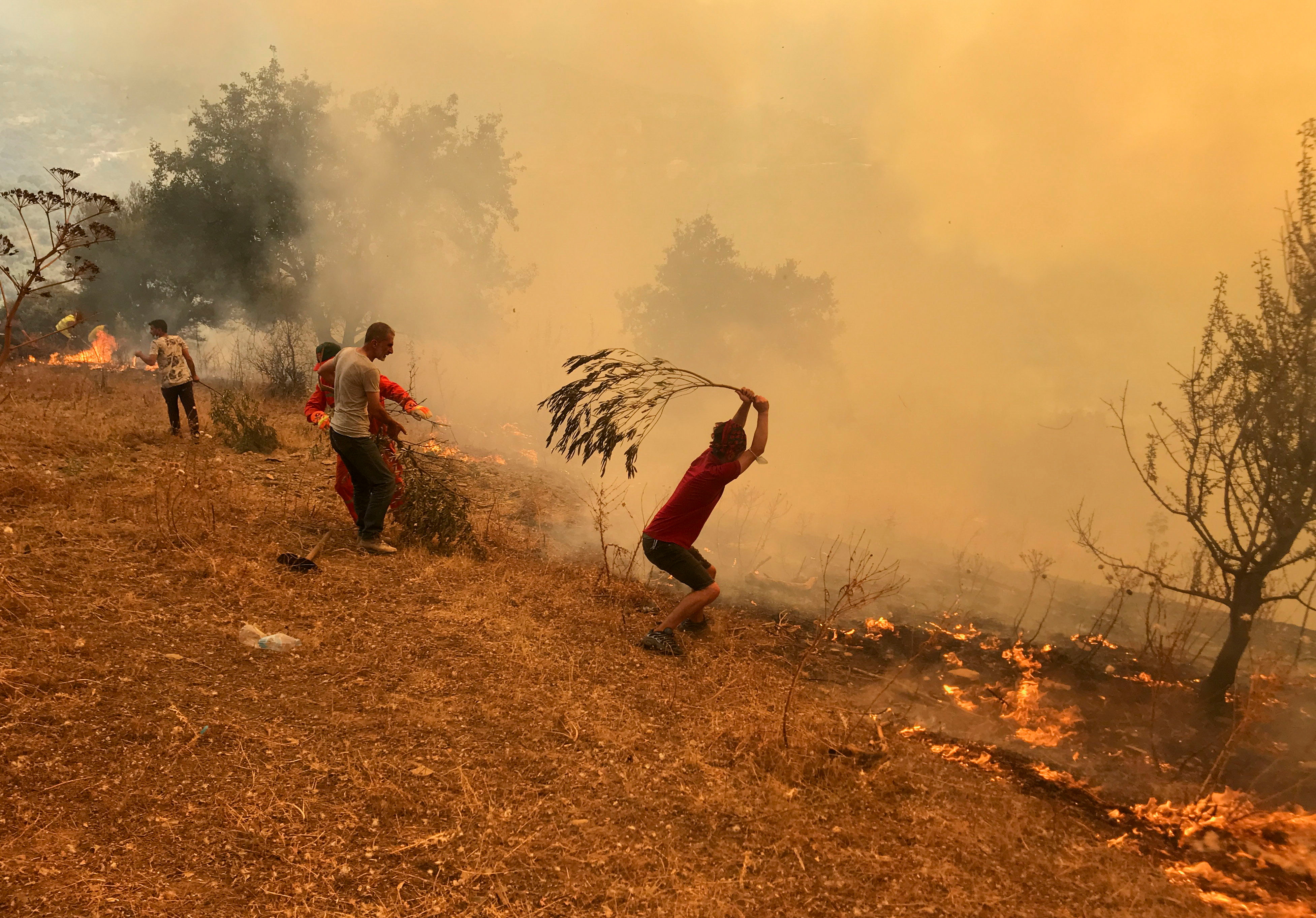 A man uses a tree branch in an attempt to ward off the wall of smoke coming at him. Everything has is tinted orange.