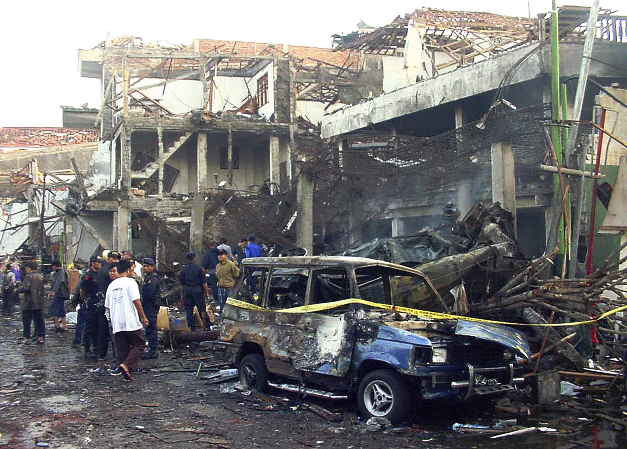The scene of the Bali bombings in 2002.. Police officers are inspecting the ruins of the devastated buildings. Others are watching. A wrecked car is in front. It has yellow police tape around it.
