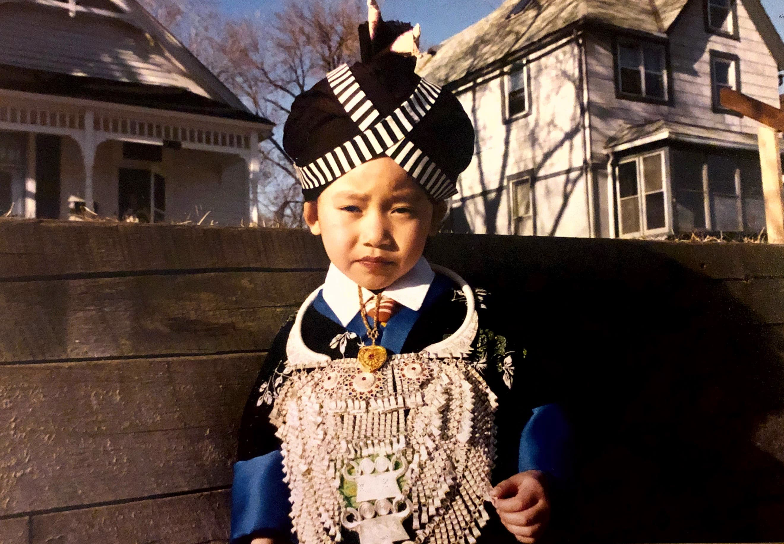 A young girl wearing a traditional Hmong dress stands in front of a house in America