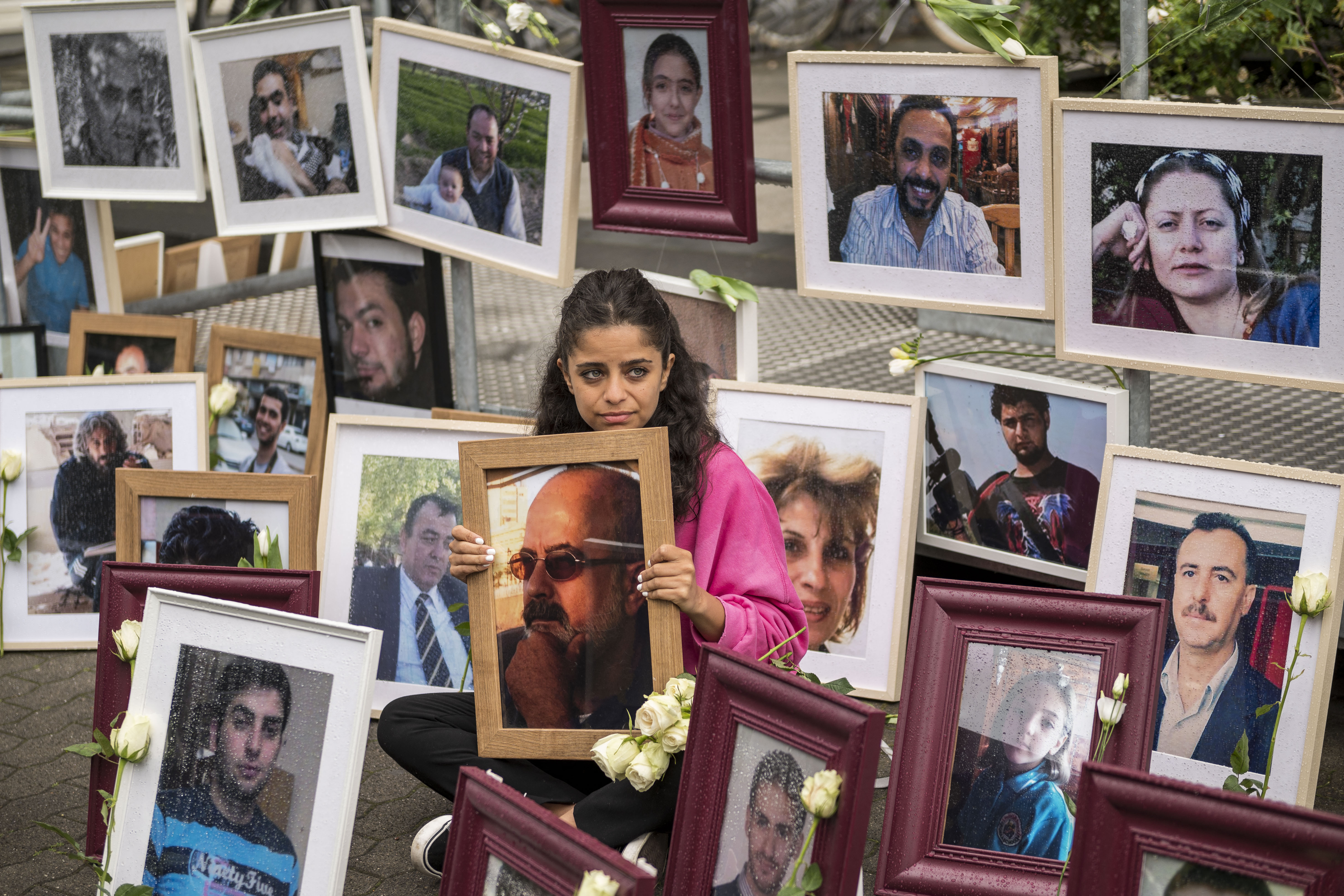 A young woman wearing a pink jumper sits on the street surrounded by framed pictures of victims of the Syrian regime.