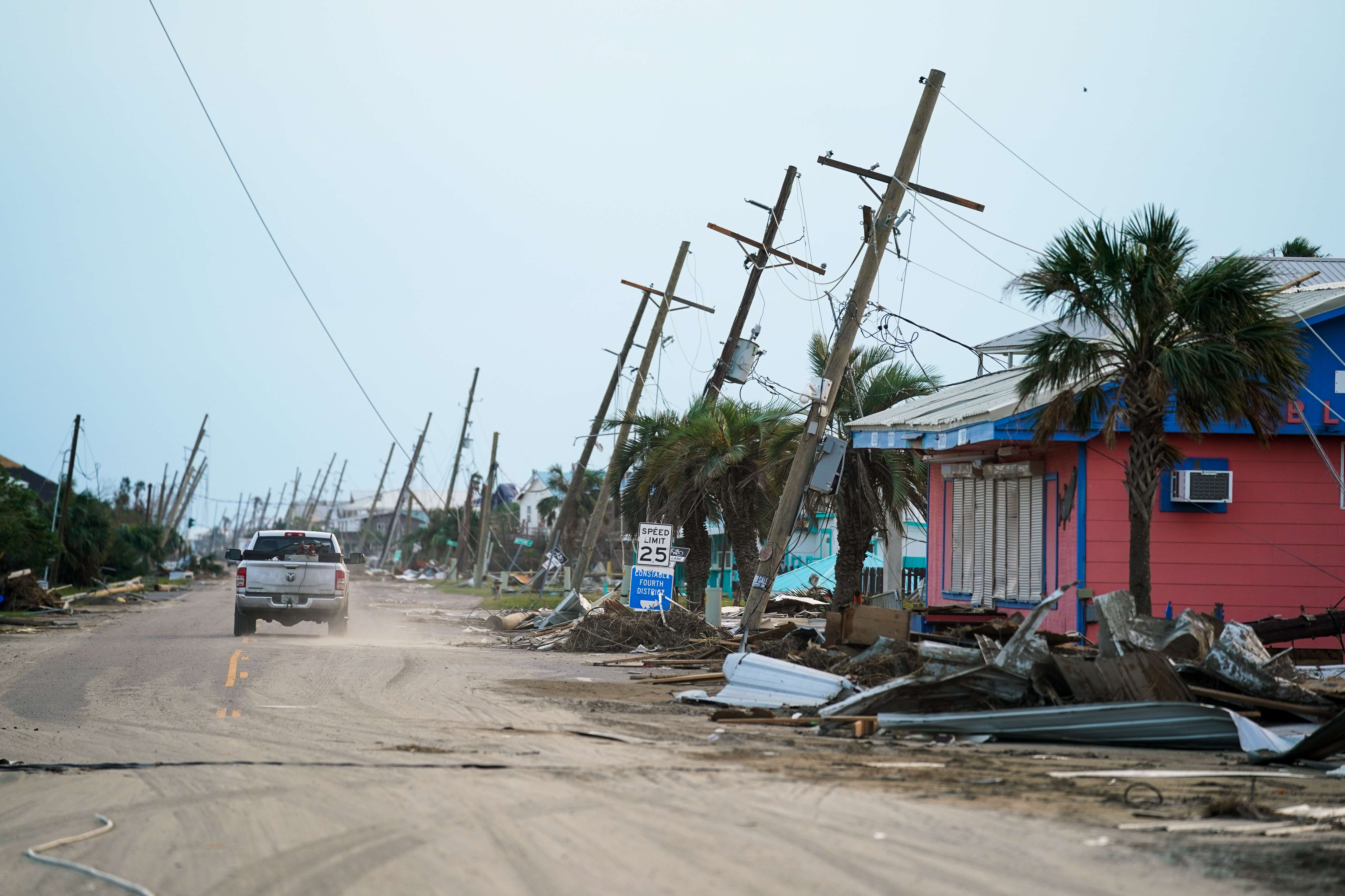A photo of a car driving down a street with a row of damaged houses and debris on the right side and a row of utility poles tilting sideways into the houses.