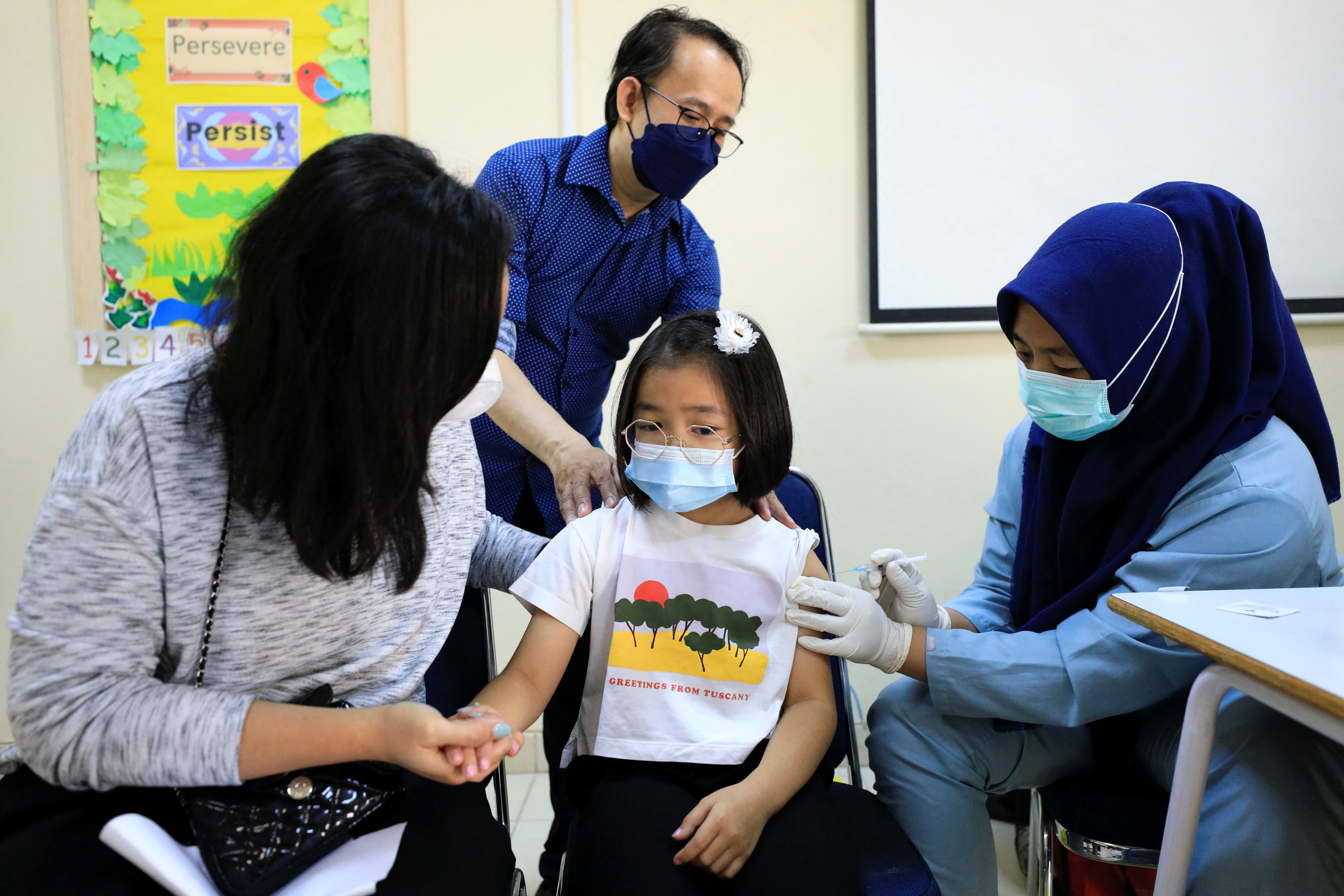 A little girl in glasses and a white t-shirt and pale blue face mask gets a COVID vaccination as her mother holds her hand
