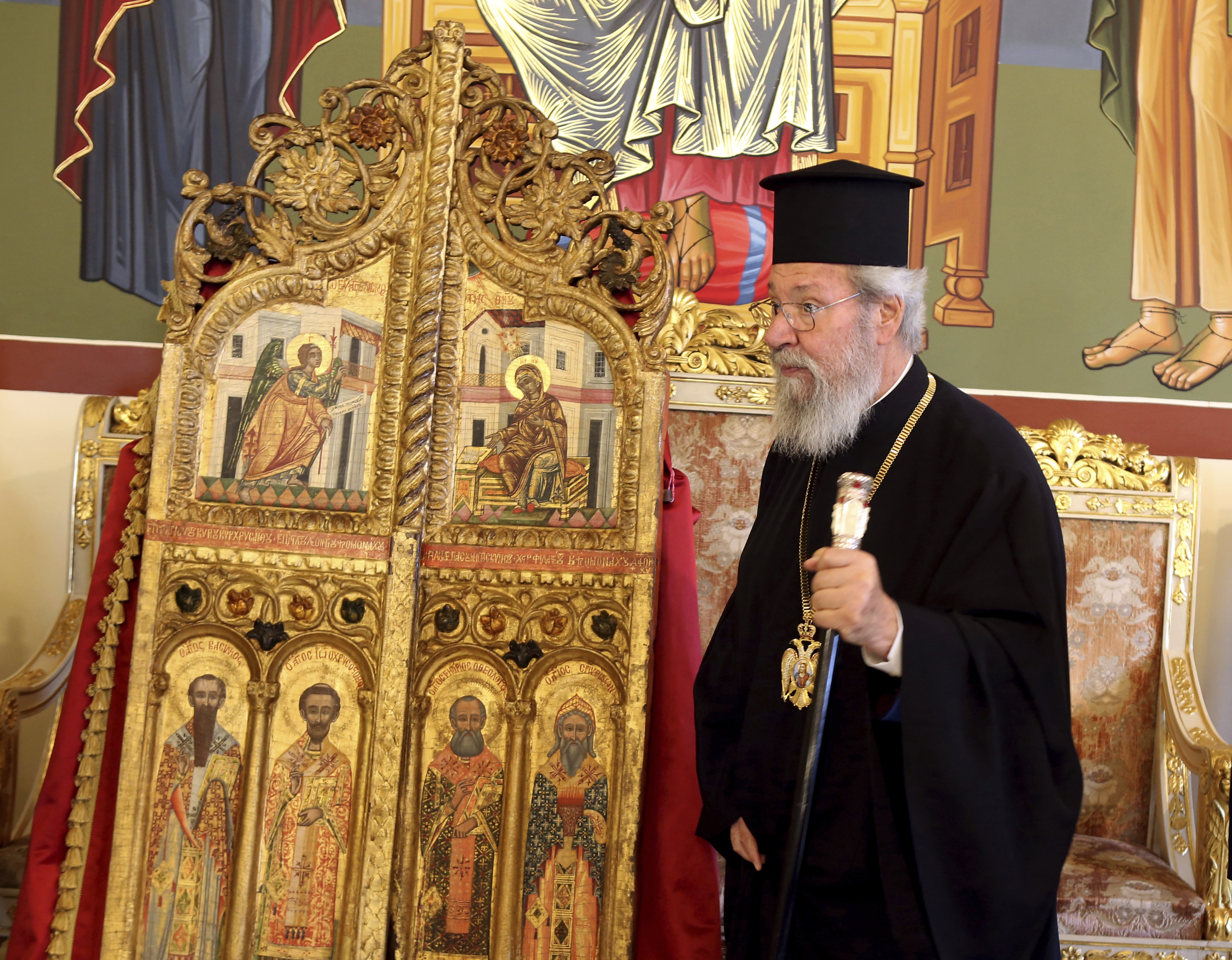 Archbishop Chrysostomos II stands next to a pair of ornate, gilded doors that guard the altar of a church