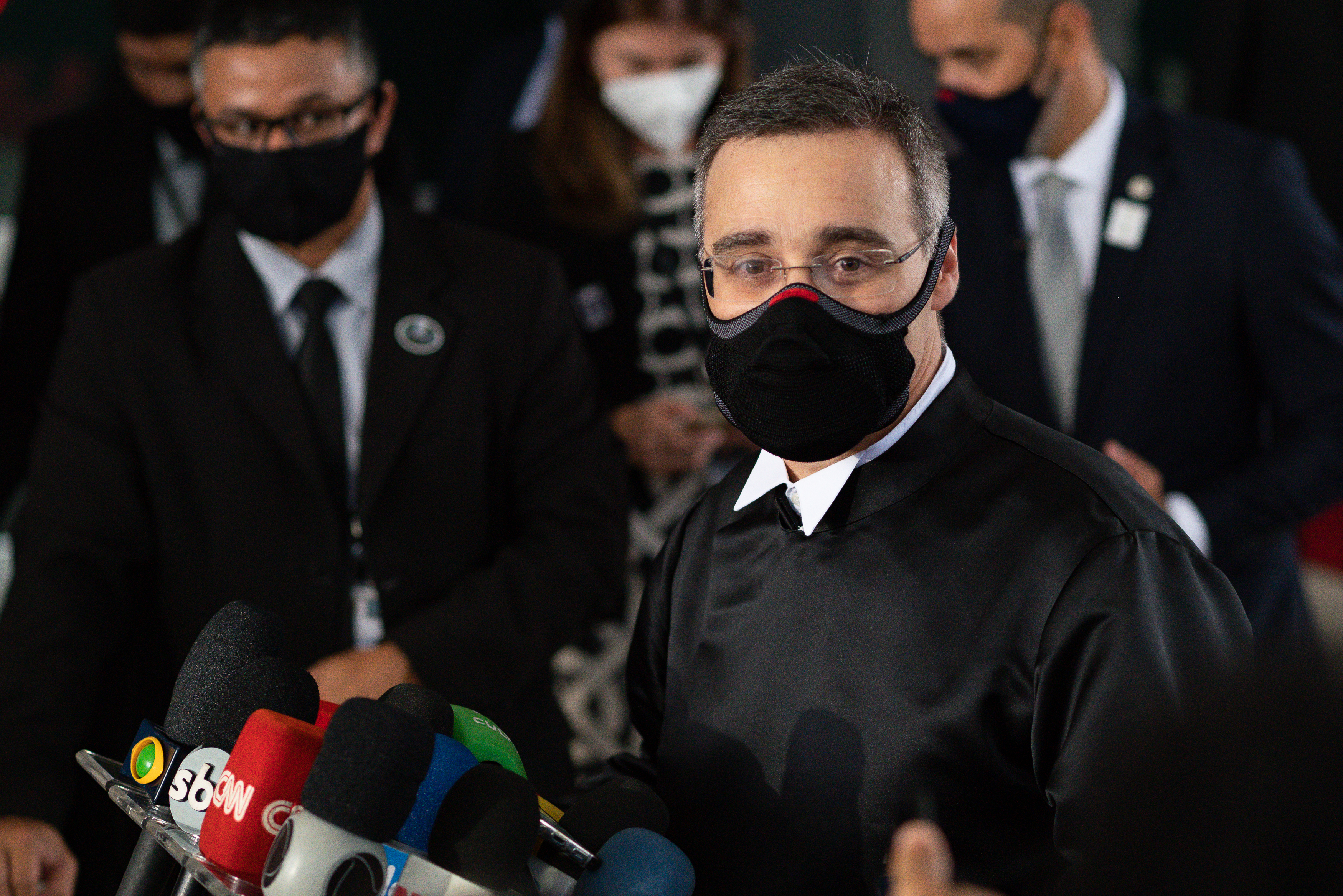 Man in black robe and a black mask talks in front of several microphones with people in suits in the background.