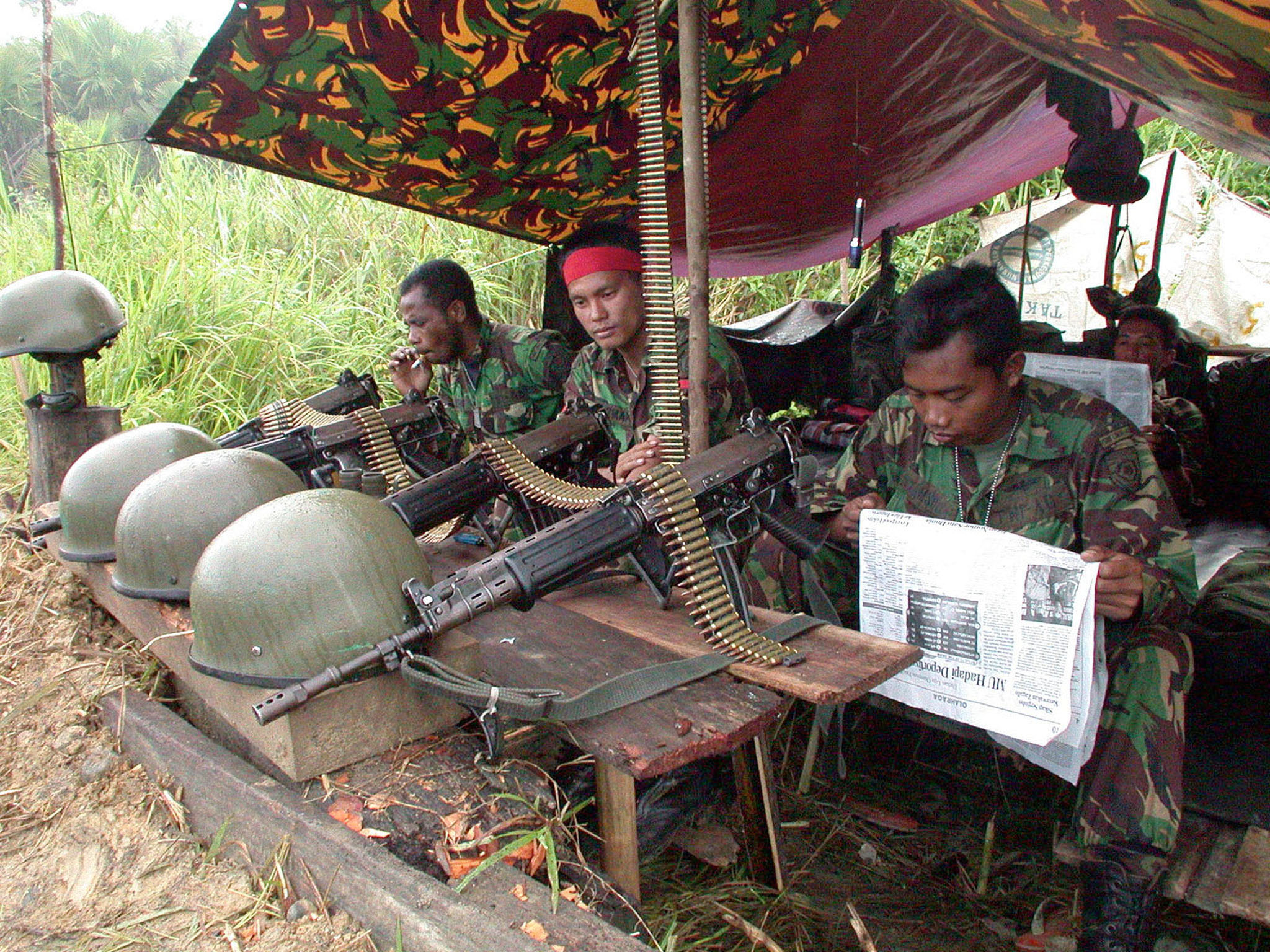 Indonesian soliders sitting under a shelter with guns and helmets on a makeshift table in front of them, in Aceh province