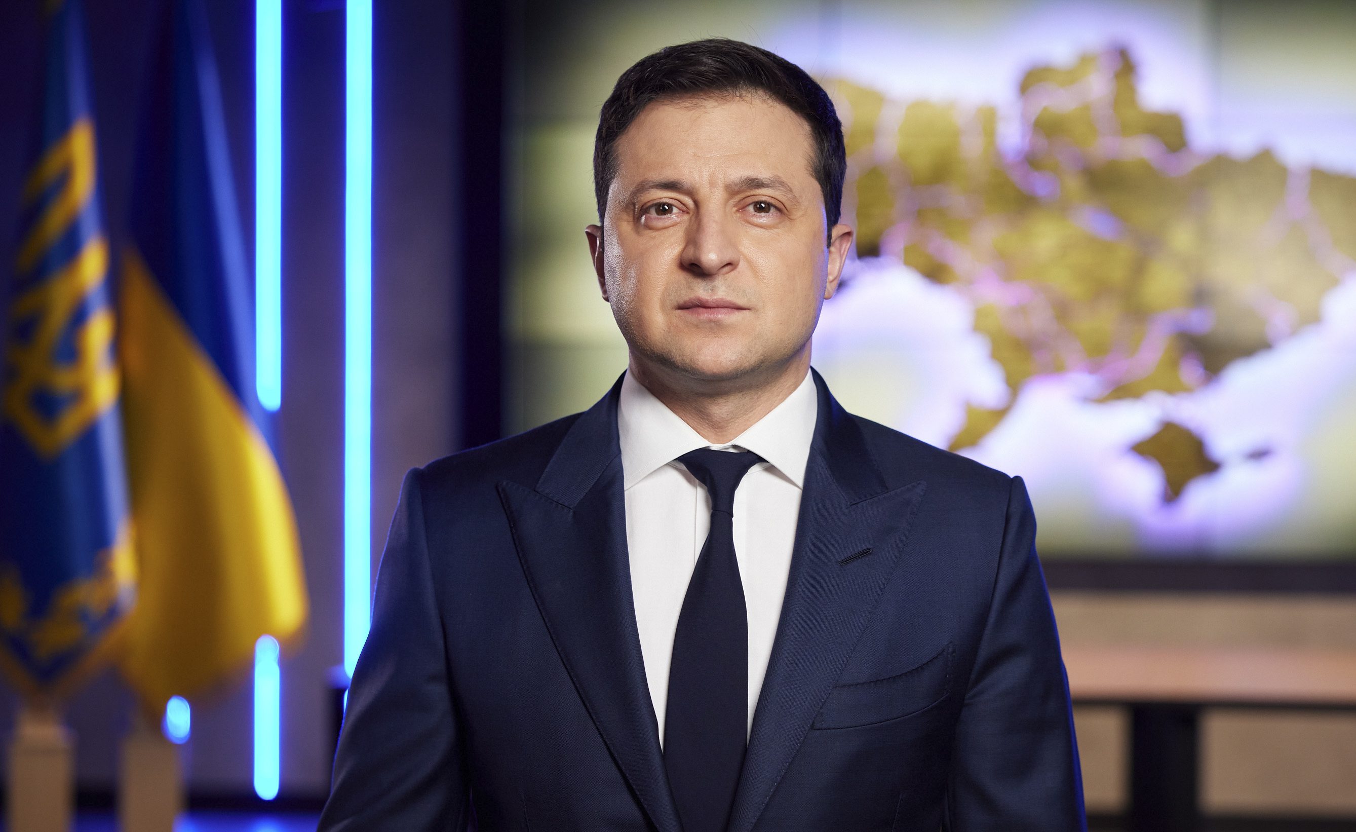 Ukrainian President Volodymyr Zelenskyy stands in front of a Ukrainian flag.