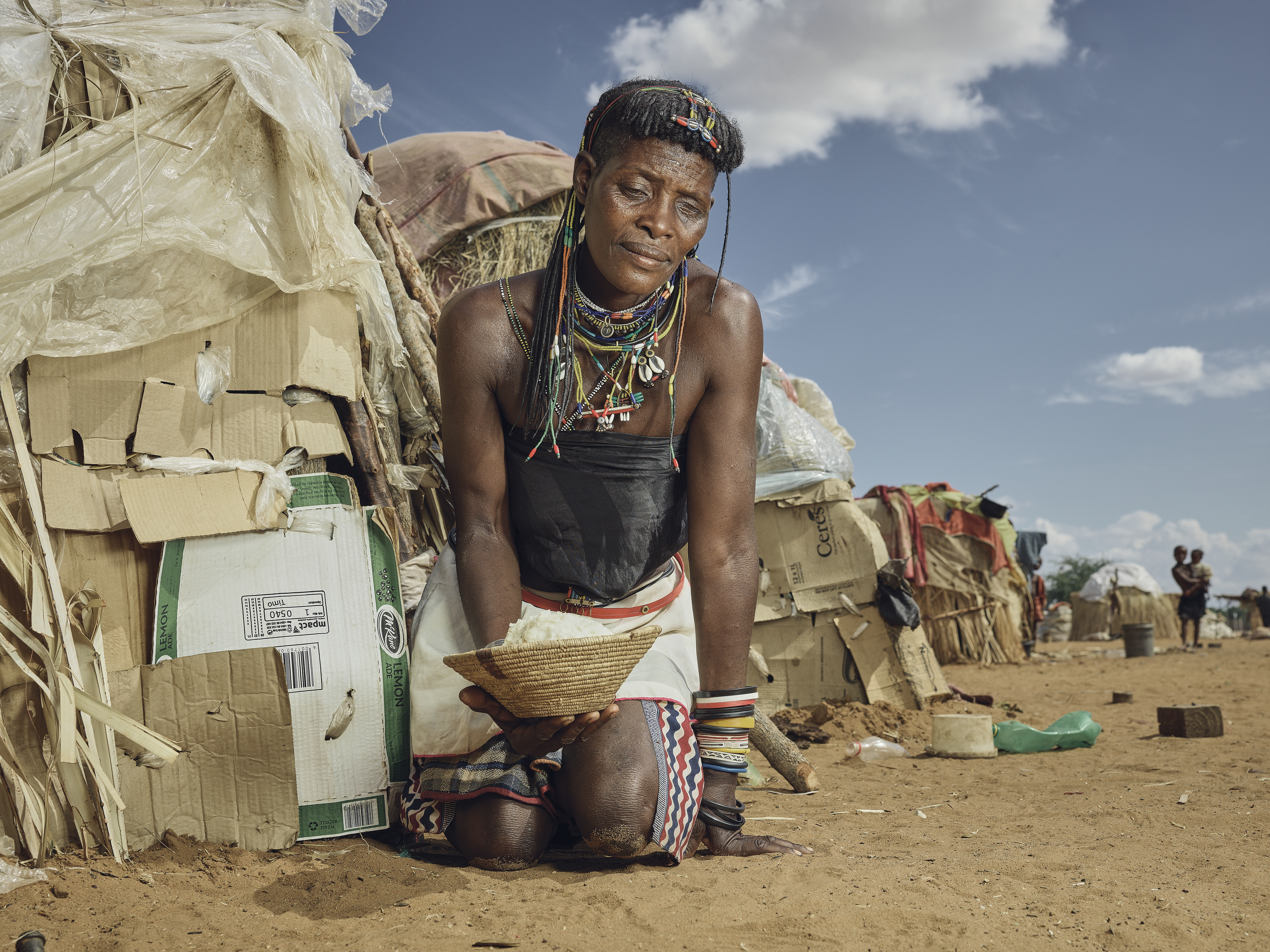 A photo of an Indigenous refugee kneeling down, holding a bowl of rice outside a refugee shelter.