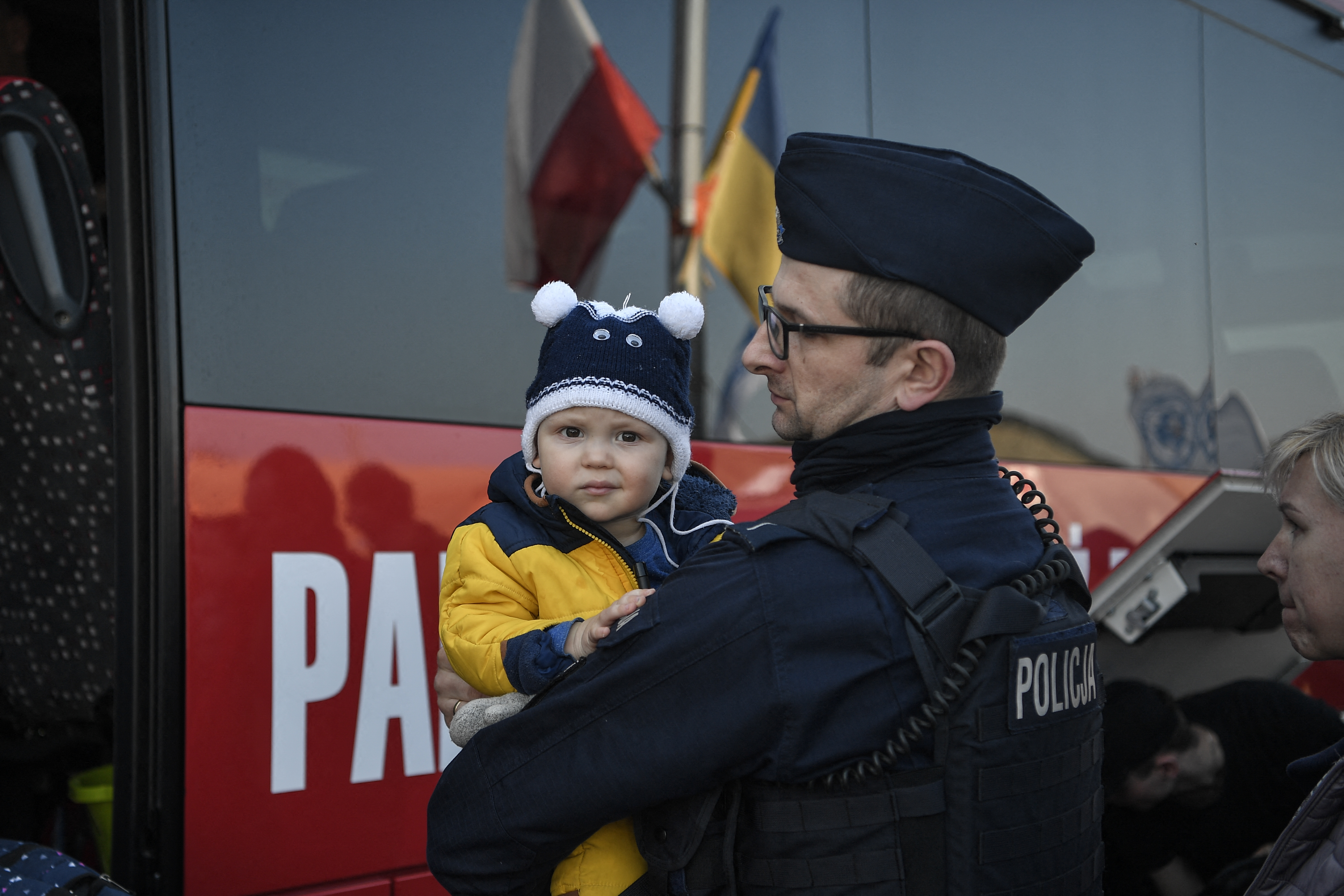 A Polish police officer holds a child to help his mother while board a bus for their further transportation after crossing from Ukraine into Poland at the Medyka border crossing on March 15, 2022. - More than three million people have now fled Ukraine since Russia invaded on February 24, the United Nations said on March 15, 2022. (Photo by Louisa GOULIAMAKI / AFP)
