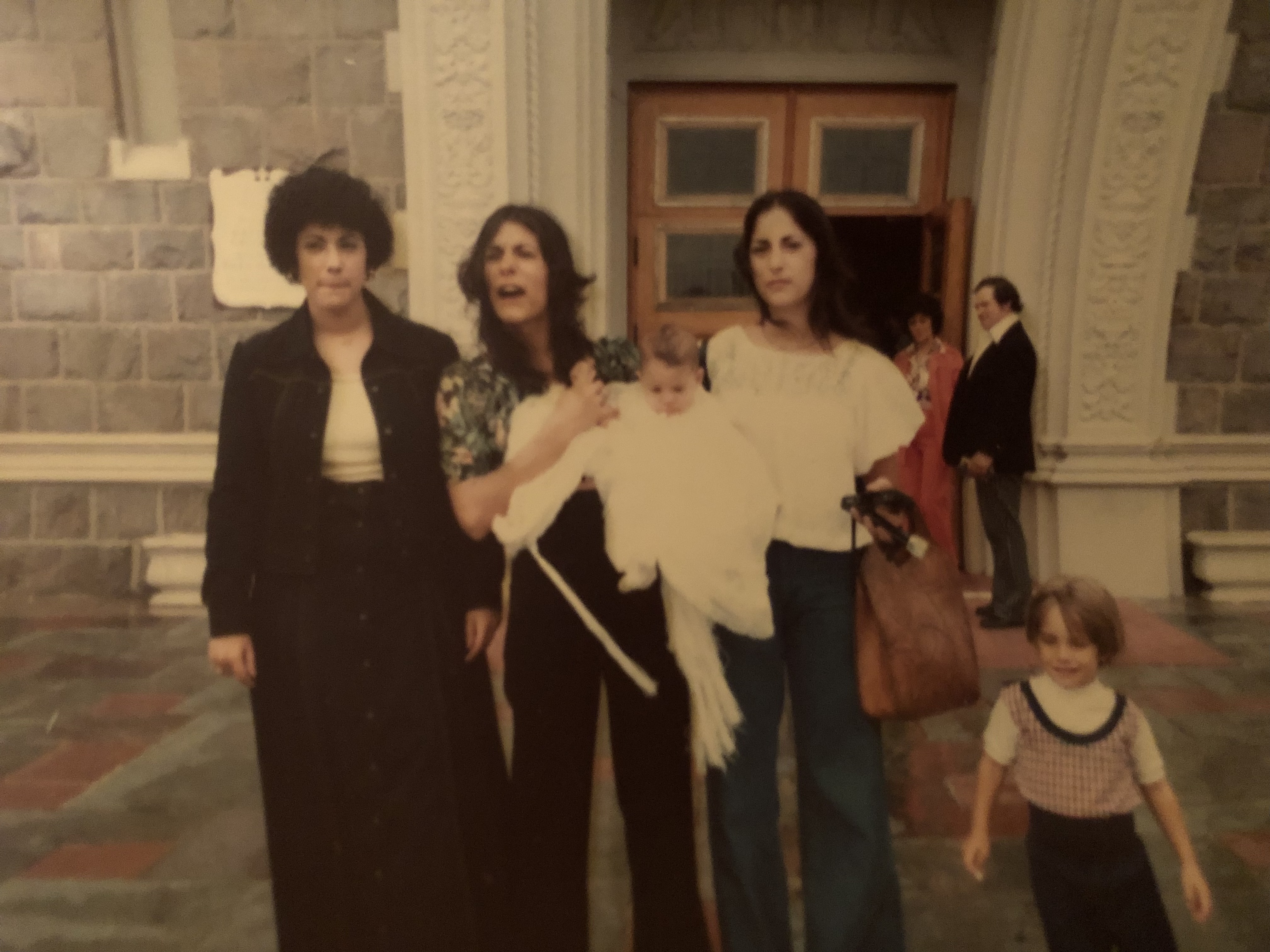 A photo of three women standing outside of a church, the woman in the middle is holding a baby with the child next to them.
