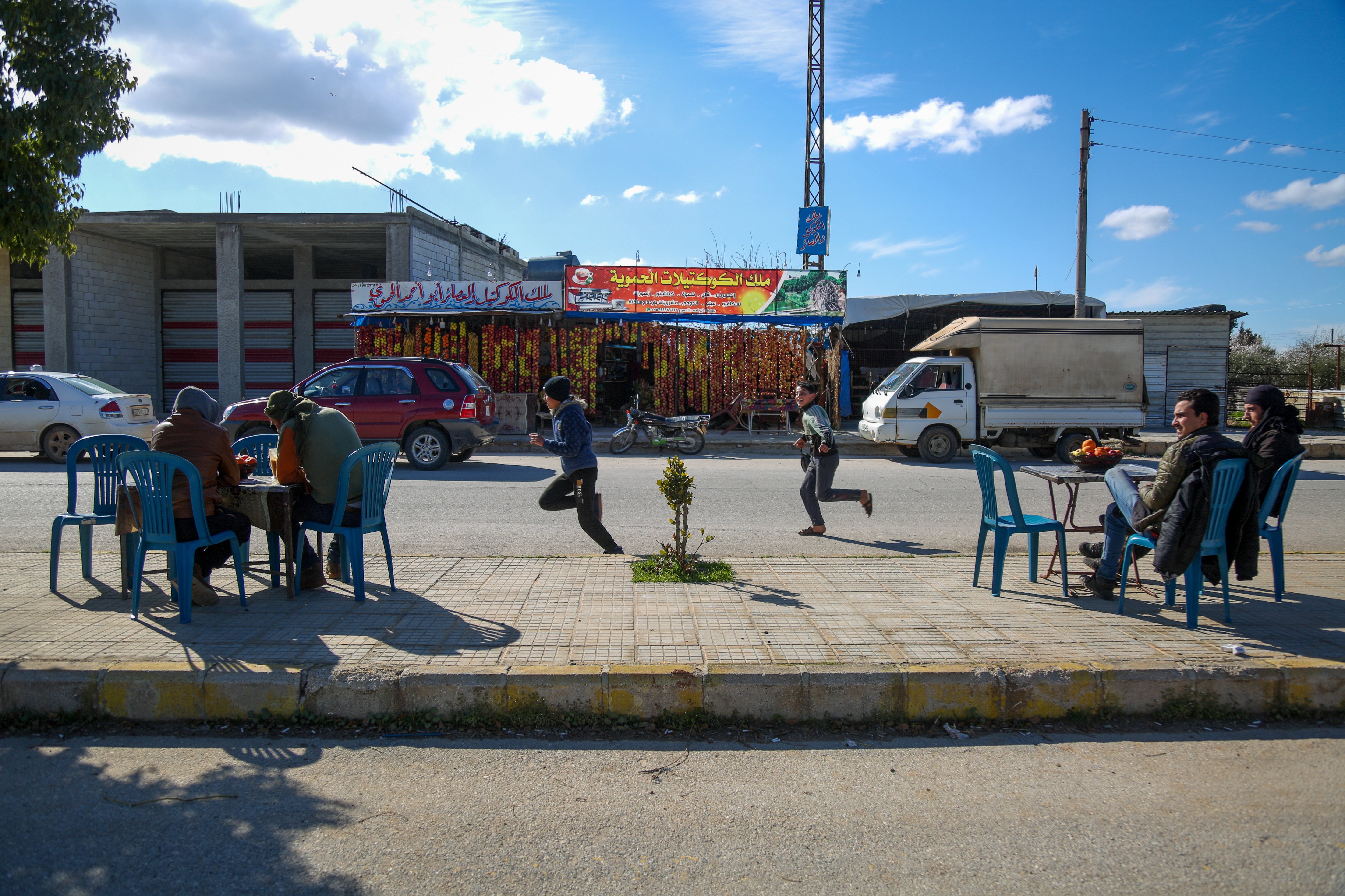 On the sidewalk opposite the juice shop, two tables of customers enjoy their drinks while children run by in the sunshine