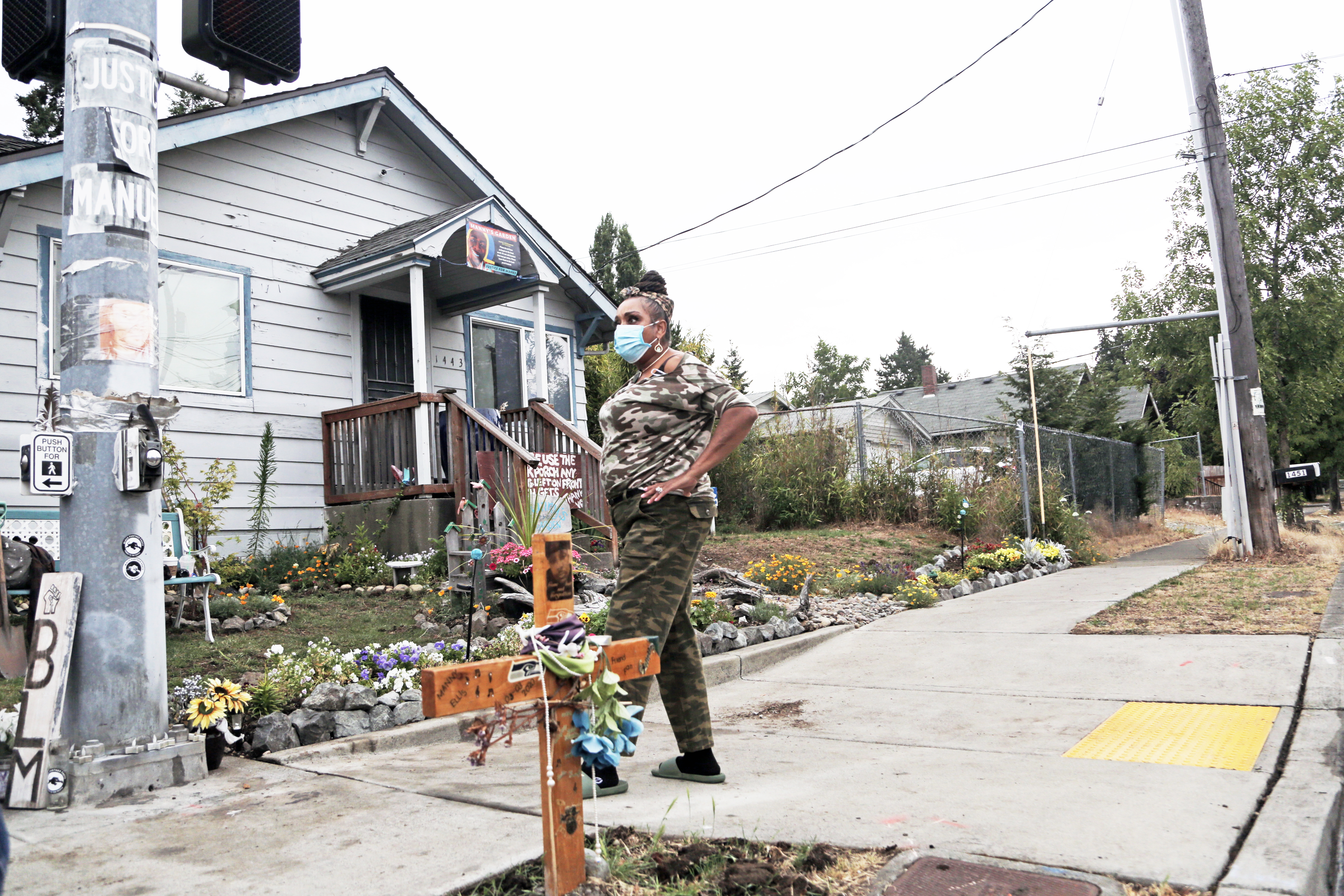 A woman stands on pavement in front of a single-storey house. Beside her, on a patch of dying grass, is small wooden cross