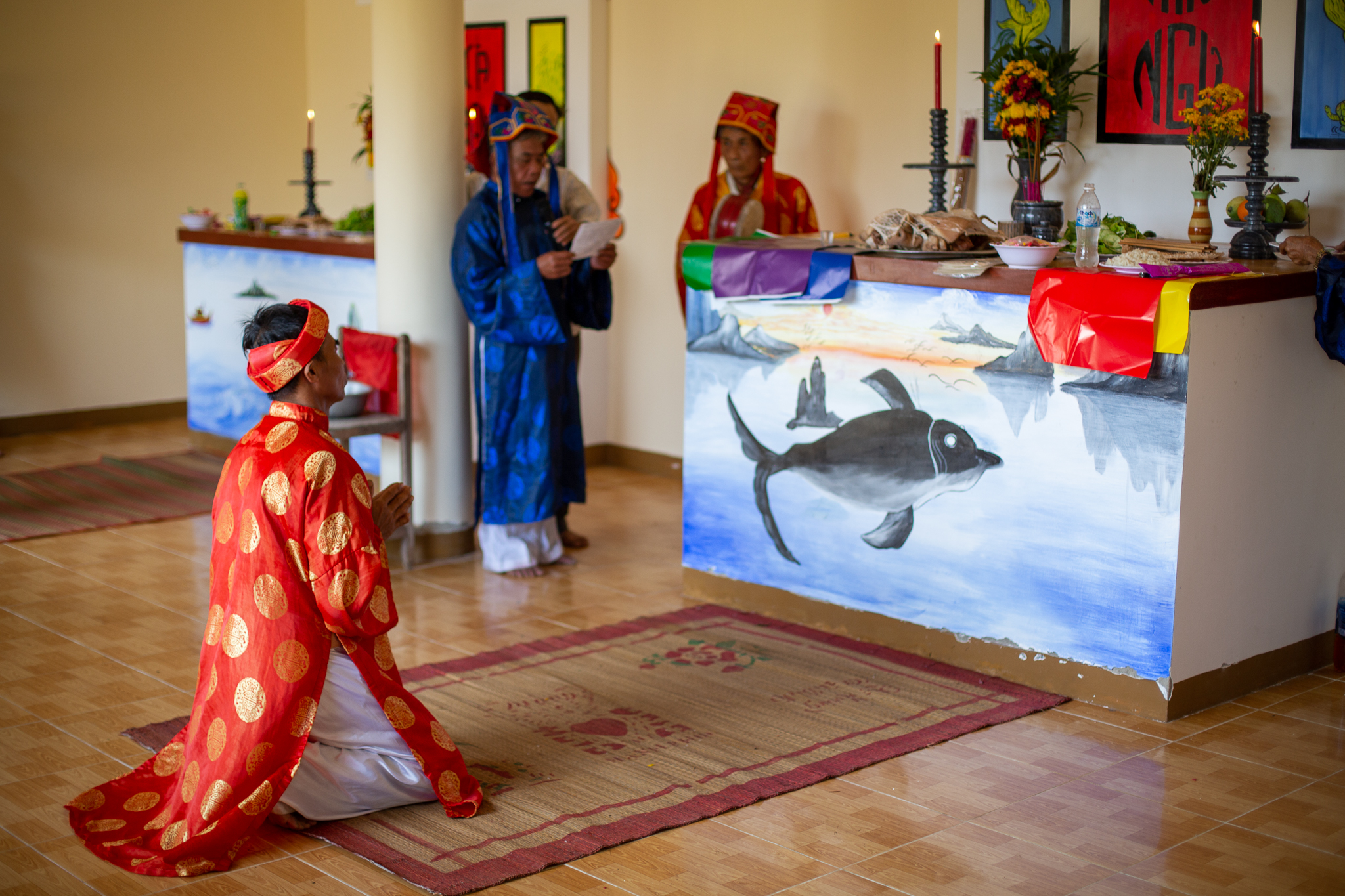 A photo of someone praying at alters where flowers, meat, fruit and rice wine rest on alters above endearing paintings of cetaceans.