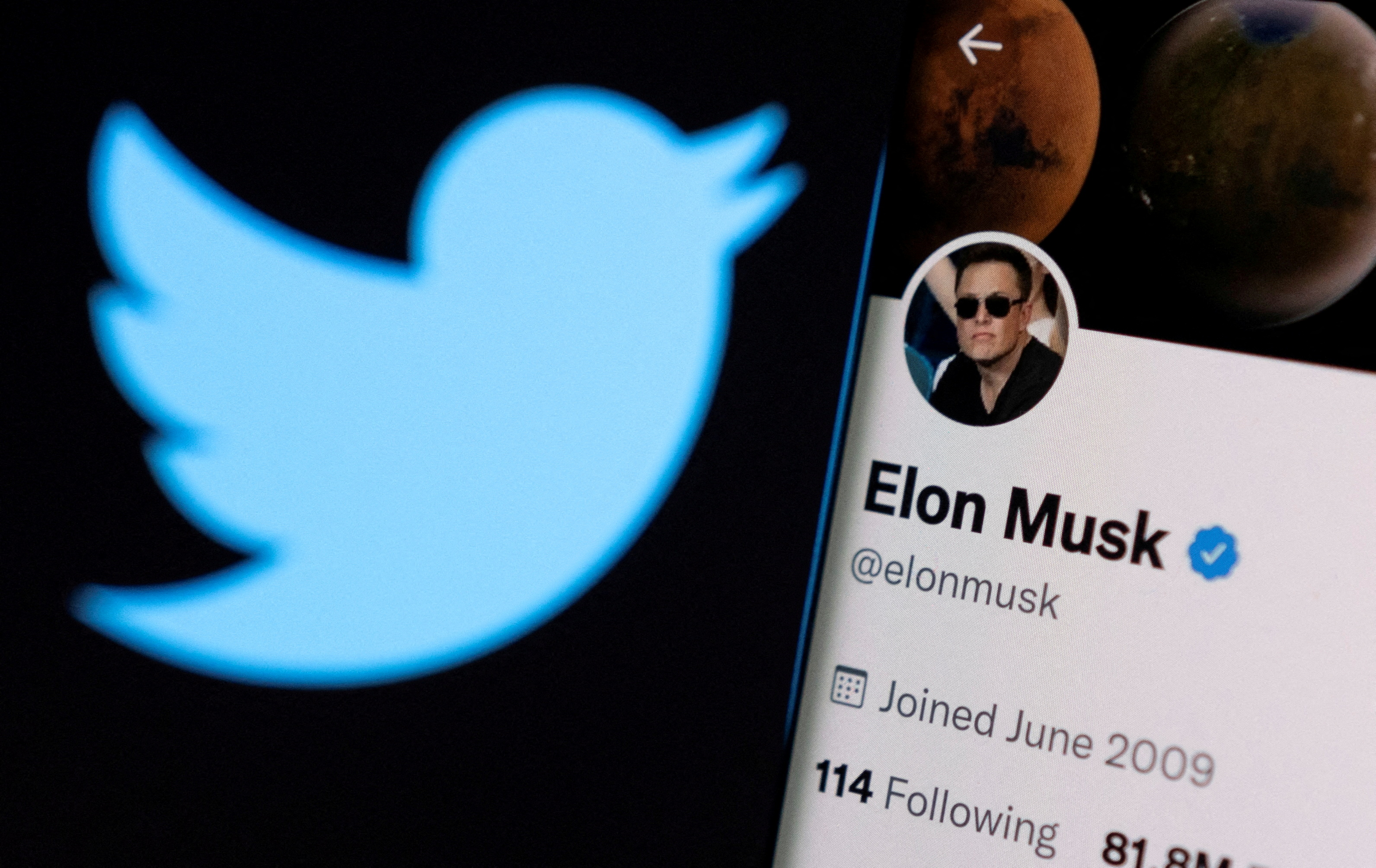 Elon Musk's twitter account is seen on a smartphone in front of the Twitter logo.