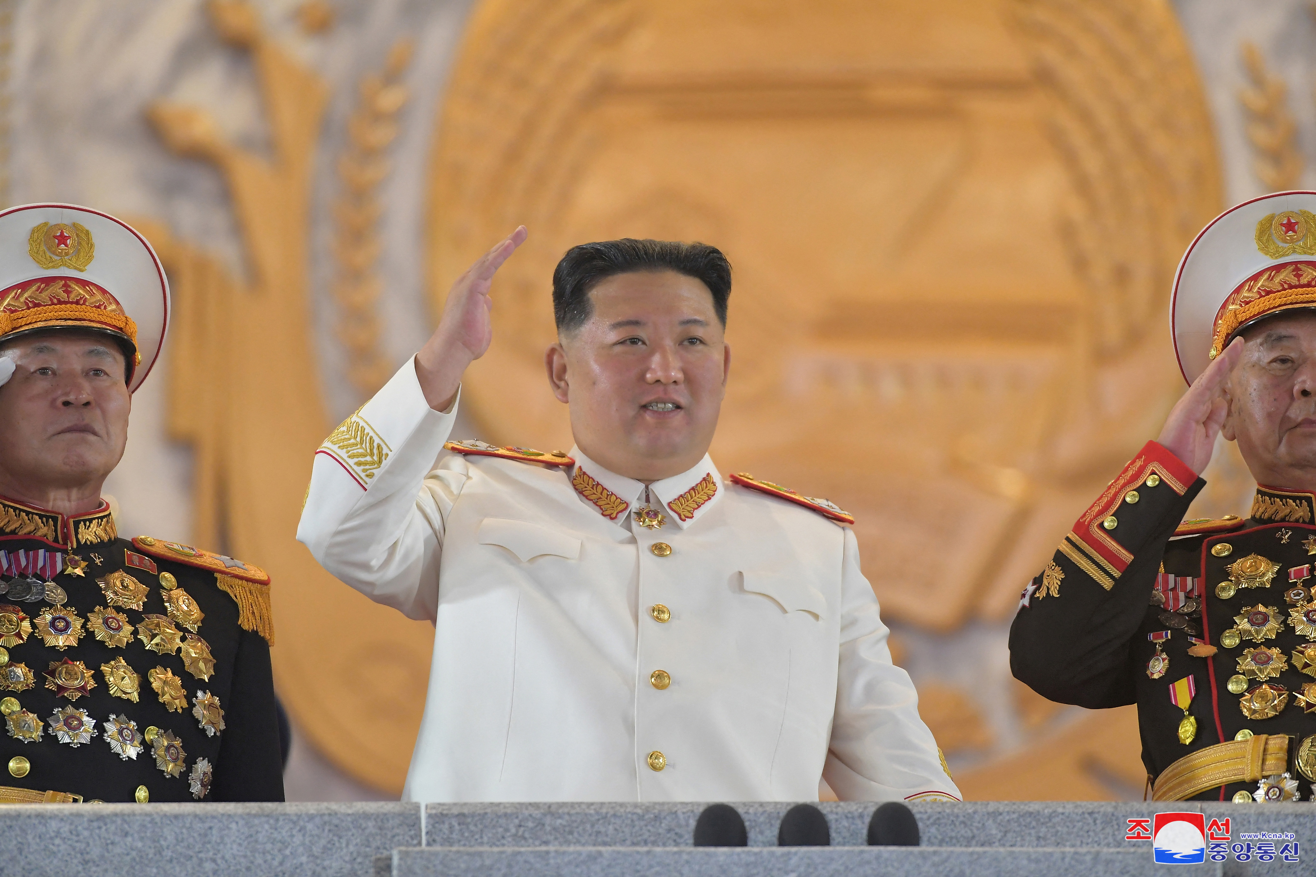 Kim Jong Un, in white ceremonial outfitm and flanked by military officers wi8th chests full of medals, acknowledges the parade from the balcony