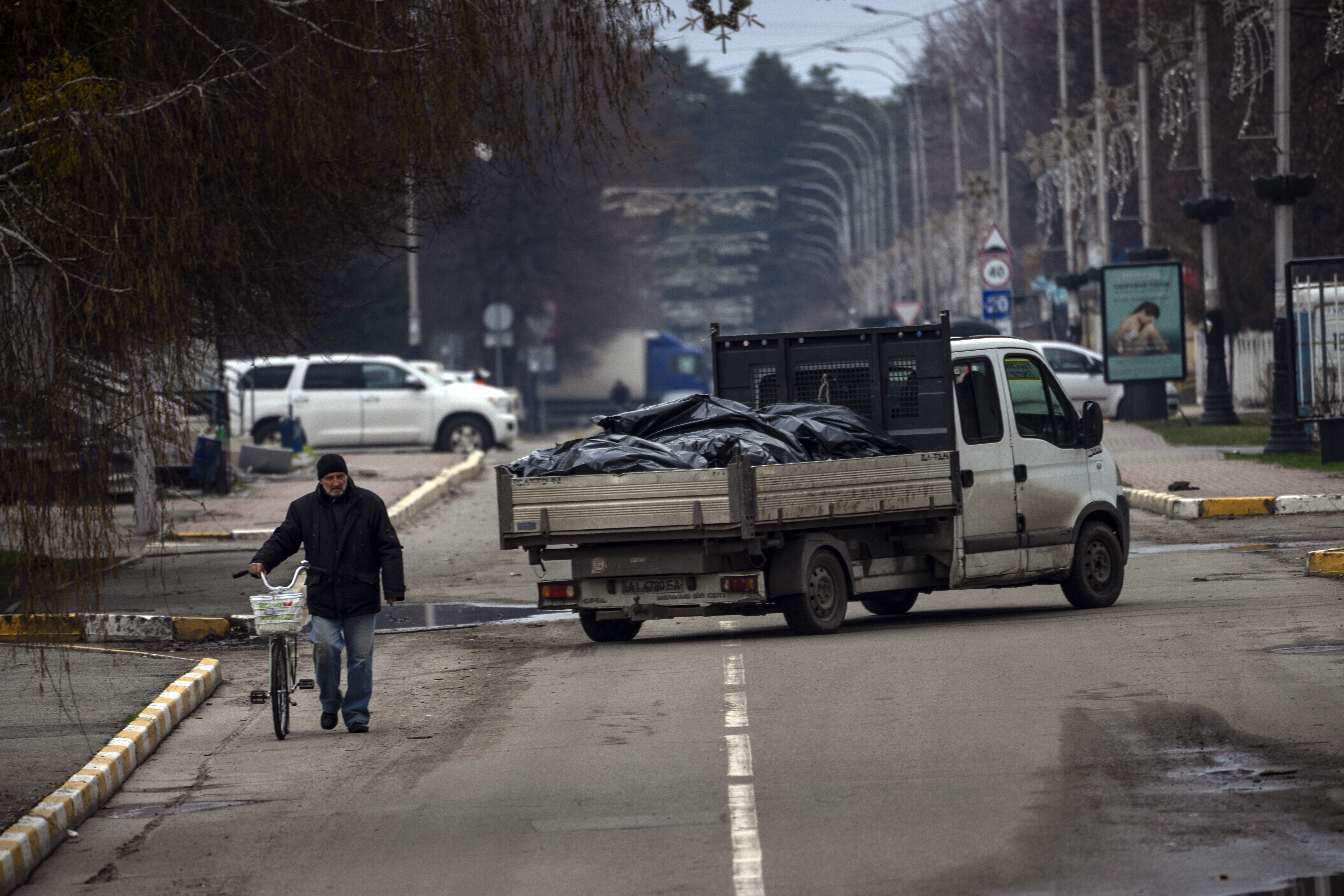 A man walks with bicycle next to truck carrying black bags with corpses exhumed from a mass grave for investigations in Bucha.