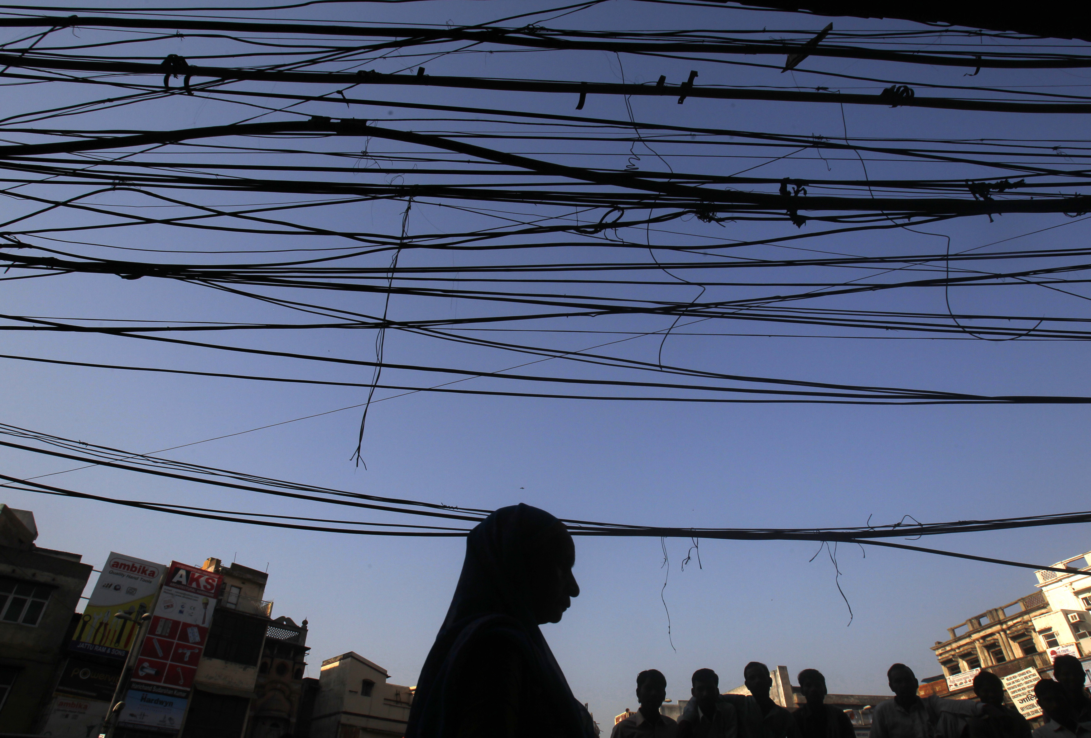 People are silhouetted against the backdrop of overhead wires and cables on a street in Delhi, India
