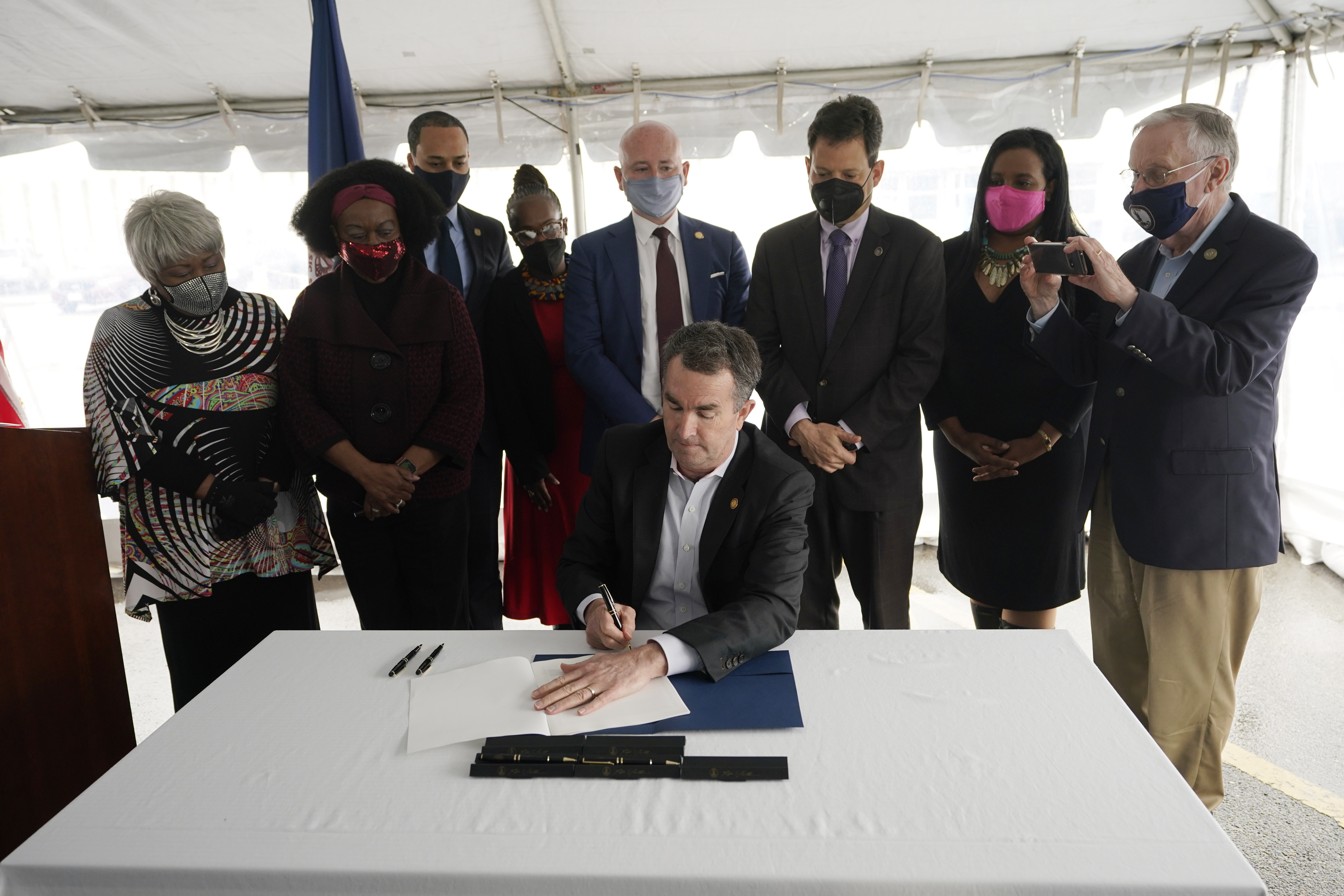 Virginia Gov. Ralph Northam seated at a table and with activists and legislators standing behind him signs the law abolishing the death penalty