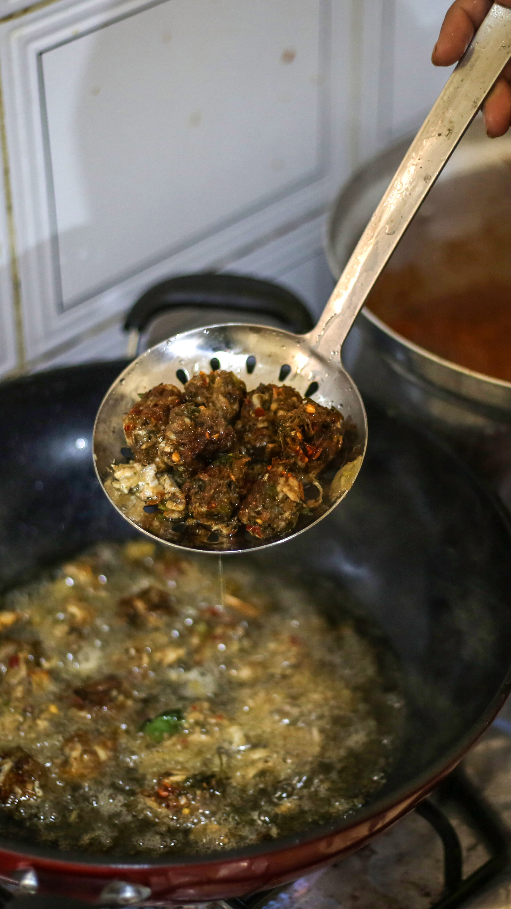 A hand holding a slotted spoon lifts some fish balls out of a wok full of bubbling oil
