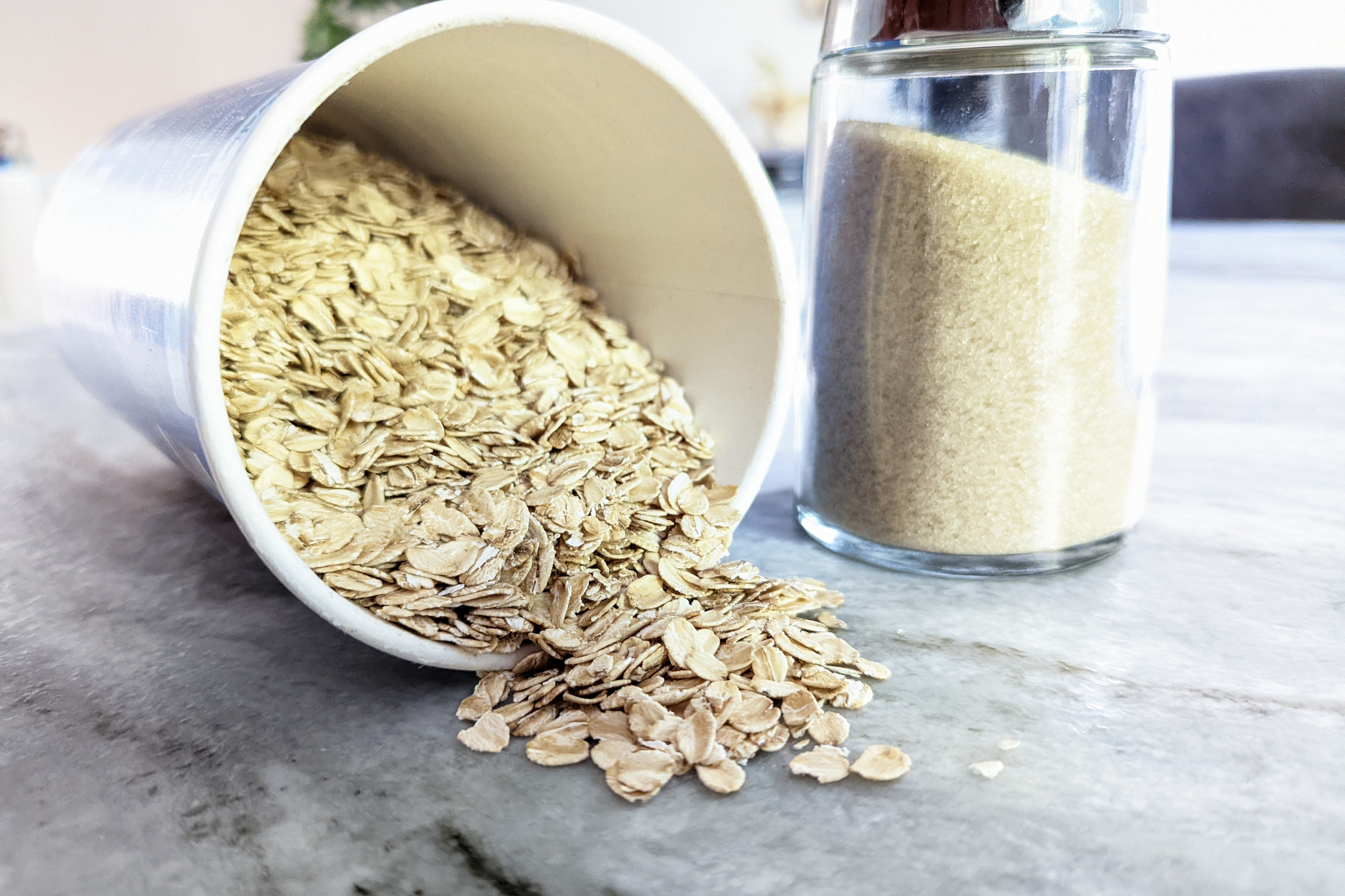 a jar of oatmeal on its side on a marble countertop, spilling some oats, standing next to it is a glass canister of sugar