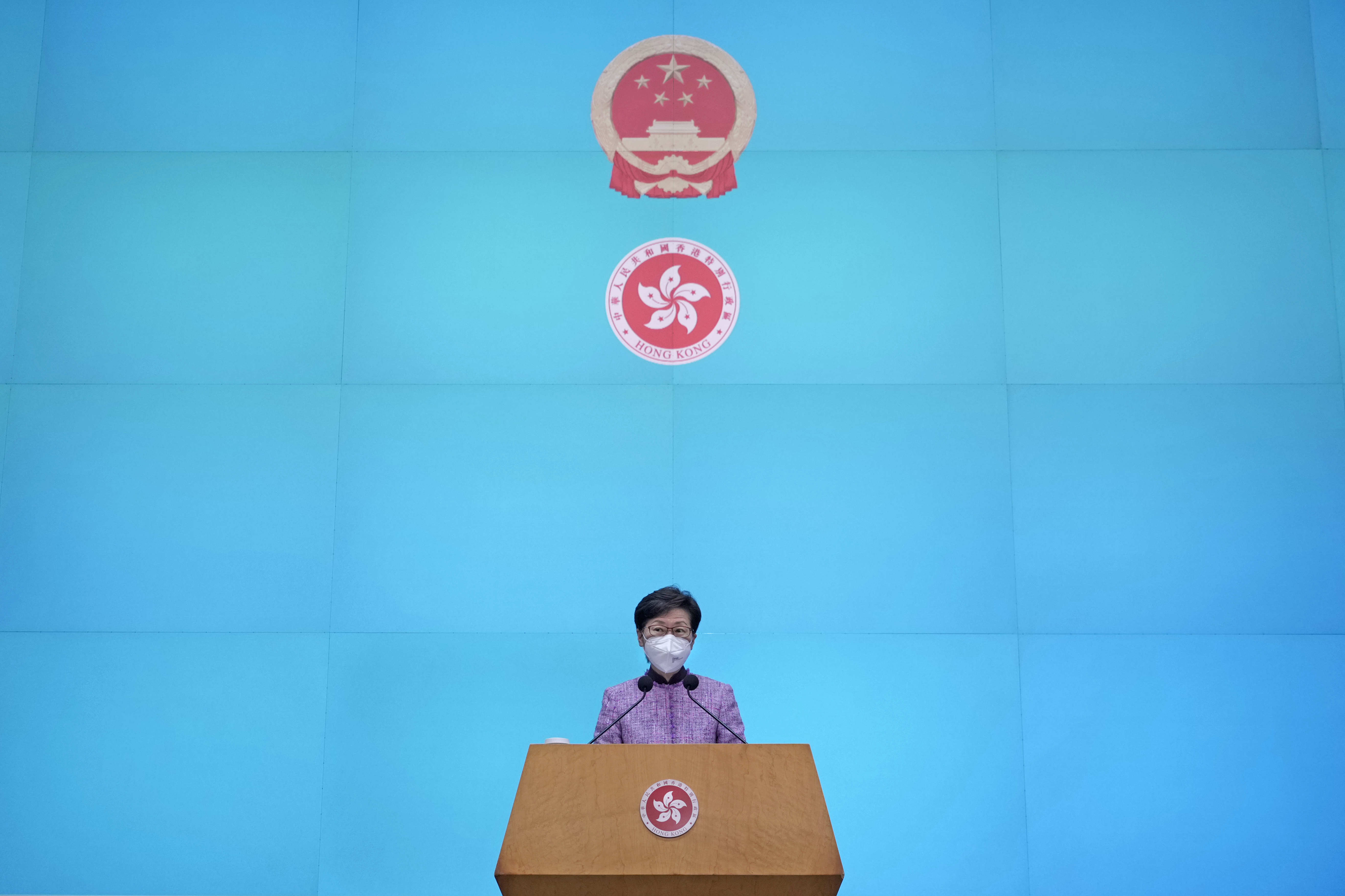 Carrle Lam stands at a lectern in front of a bright blue wall with the Hong Kong coat of arms and the Chinese Communist Party symbol behind her