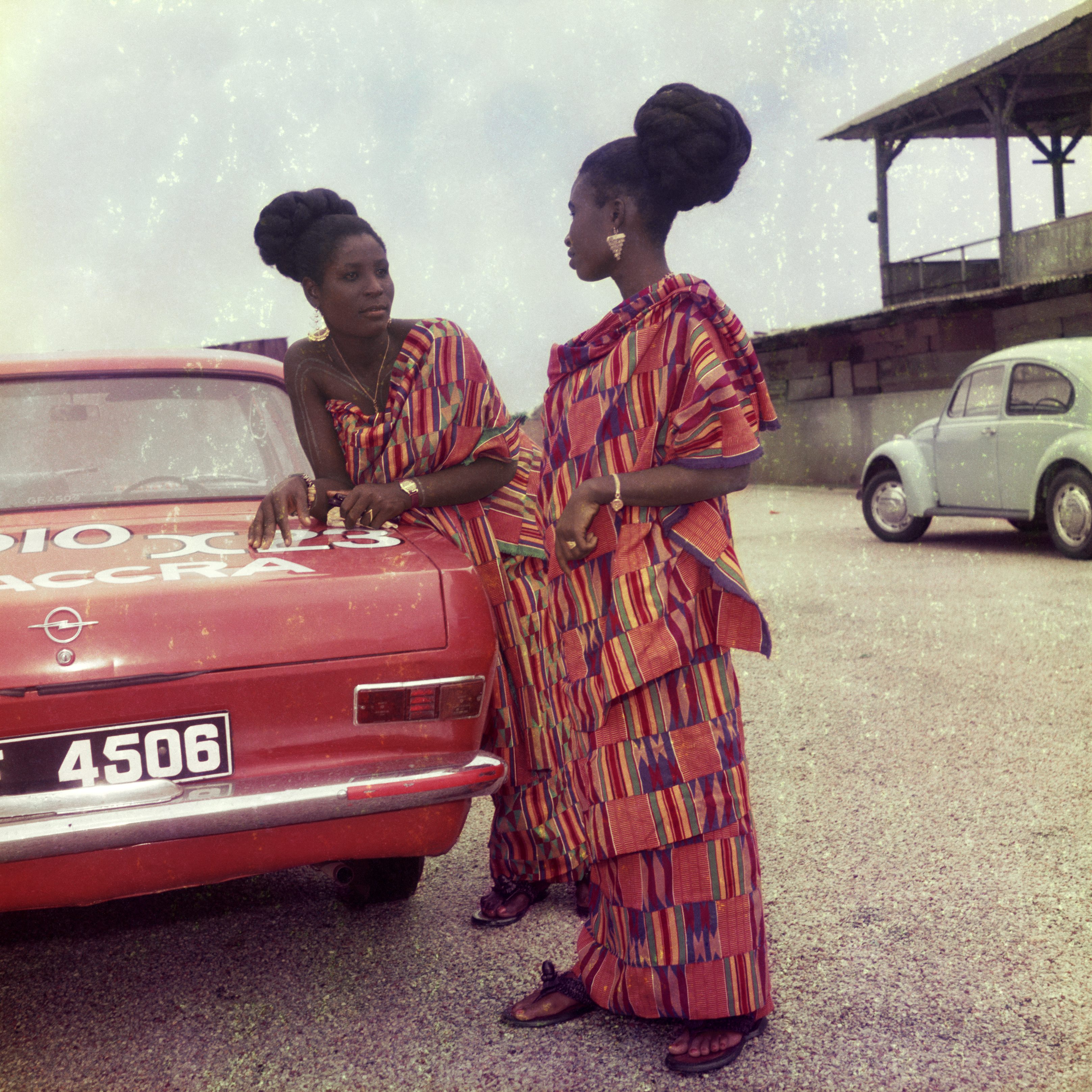 Two women dressed for a church celebration stand next to a red car