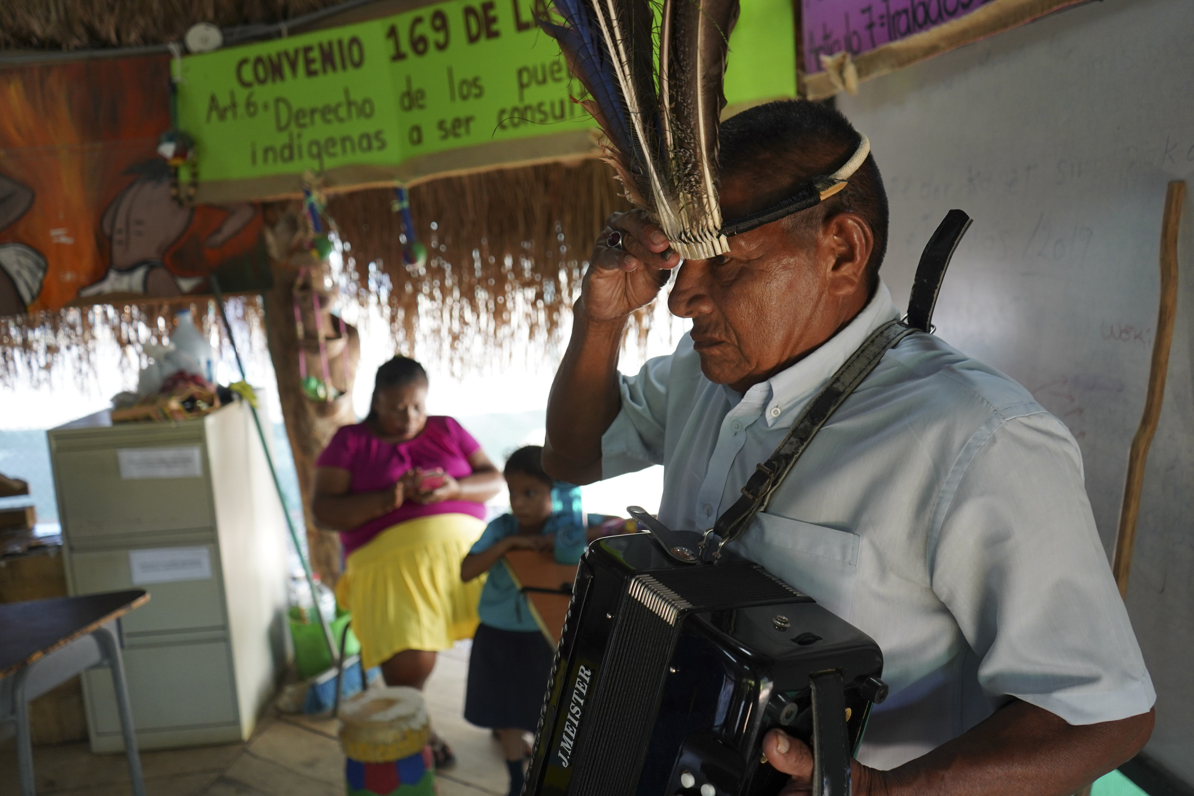 A photo of a man holding an accordion with one hand and a traditional headdress on his head with the other.