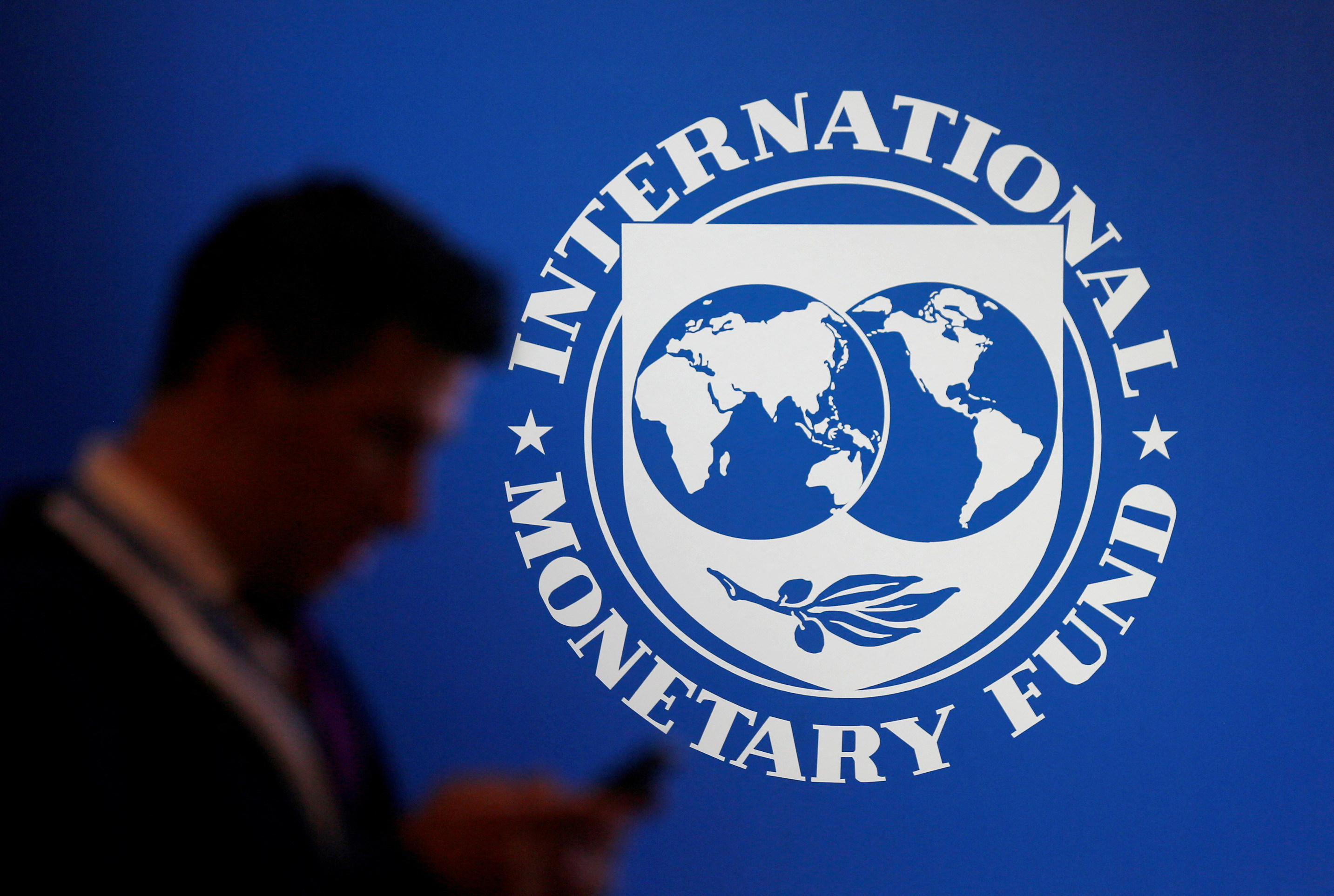 IMF logo on a wall with the silhouette of a man looking at his phone in the foreground.