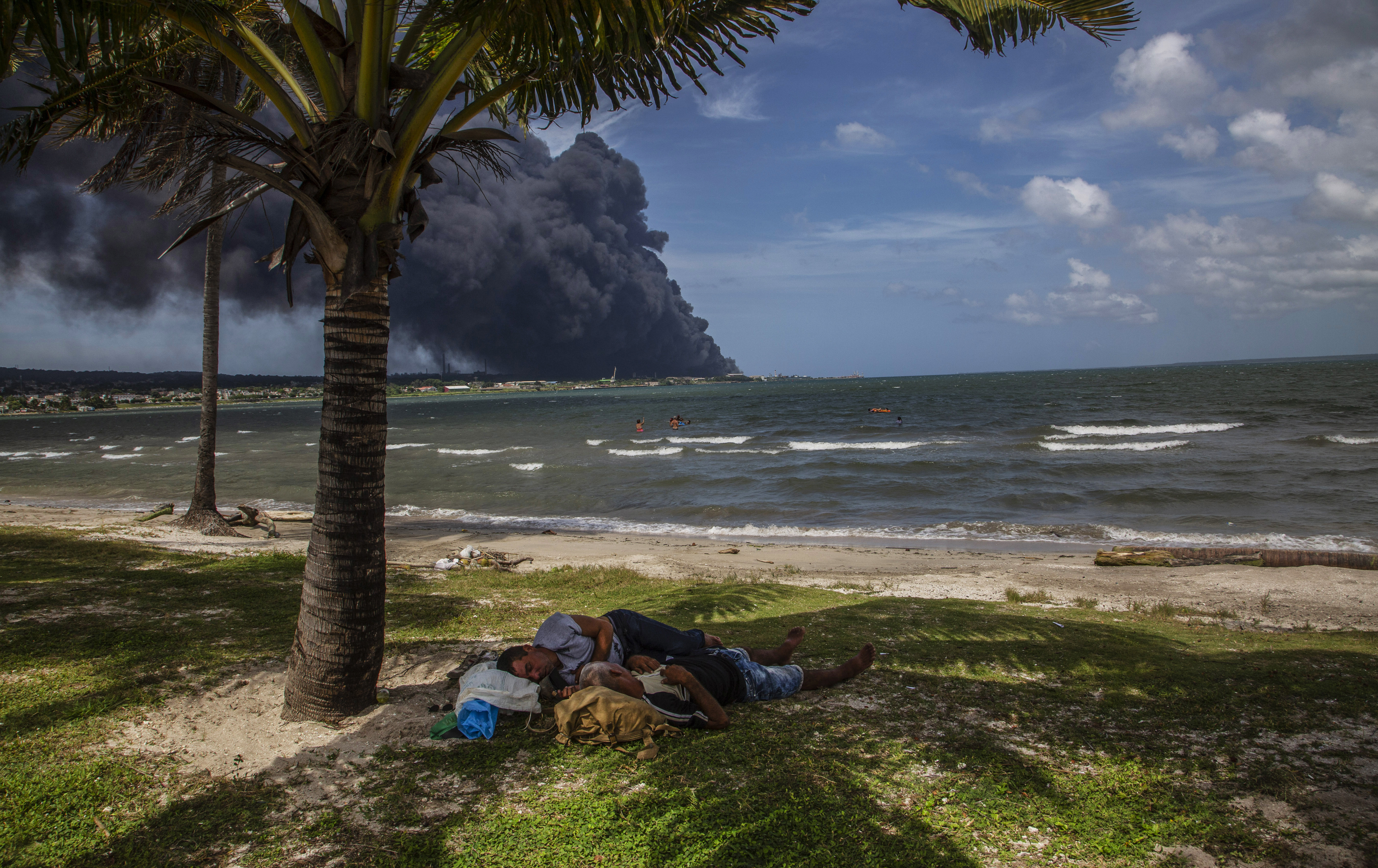 Two men sleep next to a palm tree while others take a bath near a huge plume of smoke rising from the Matanzas Supertanker Base as firefighters work to quell the blaze which began during a thunderstorm in Matanzas, Cuba