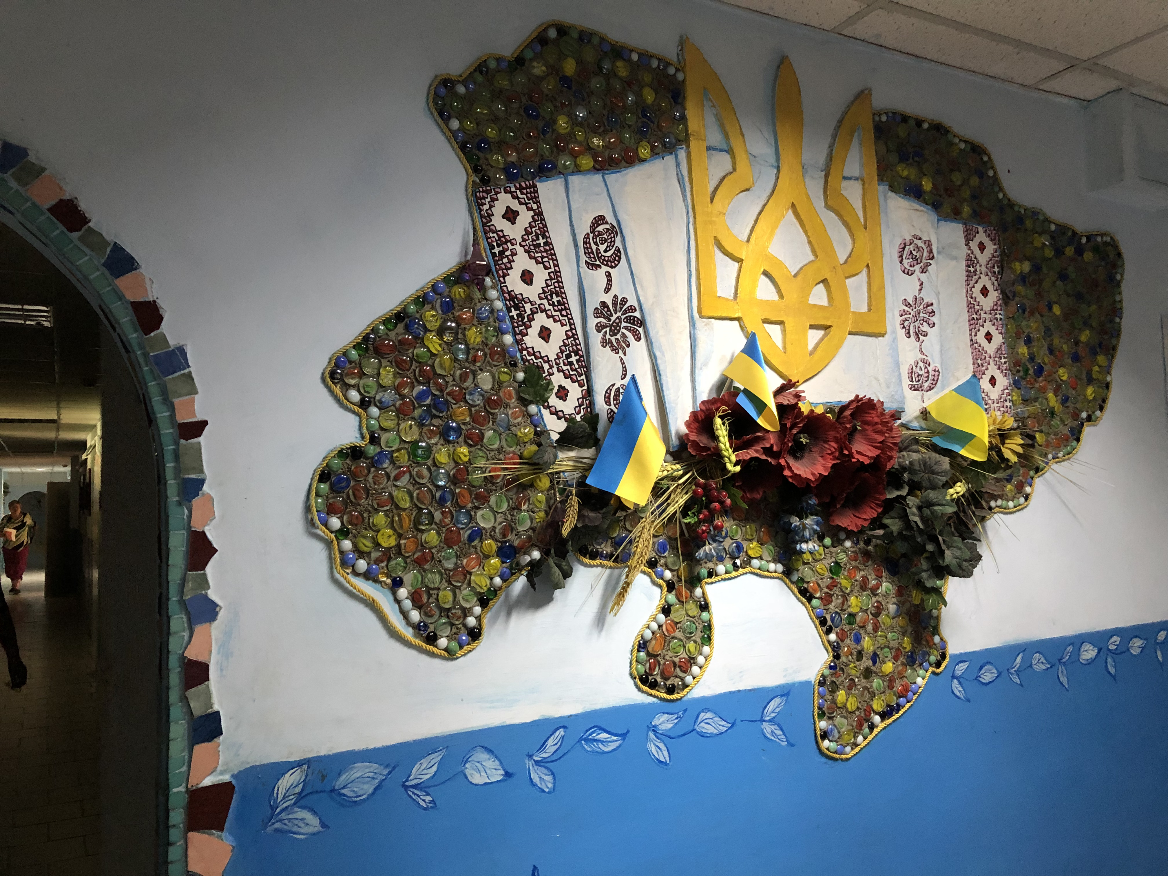 A photo of a mosaic on a wall with a few small Ukrainian flags in the middle with some flowers.