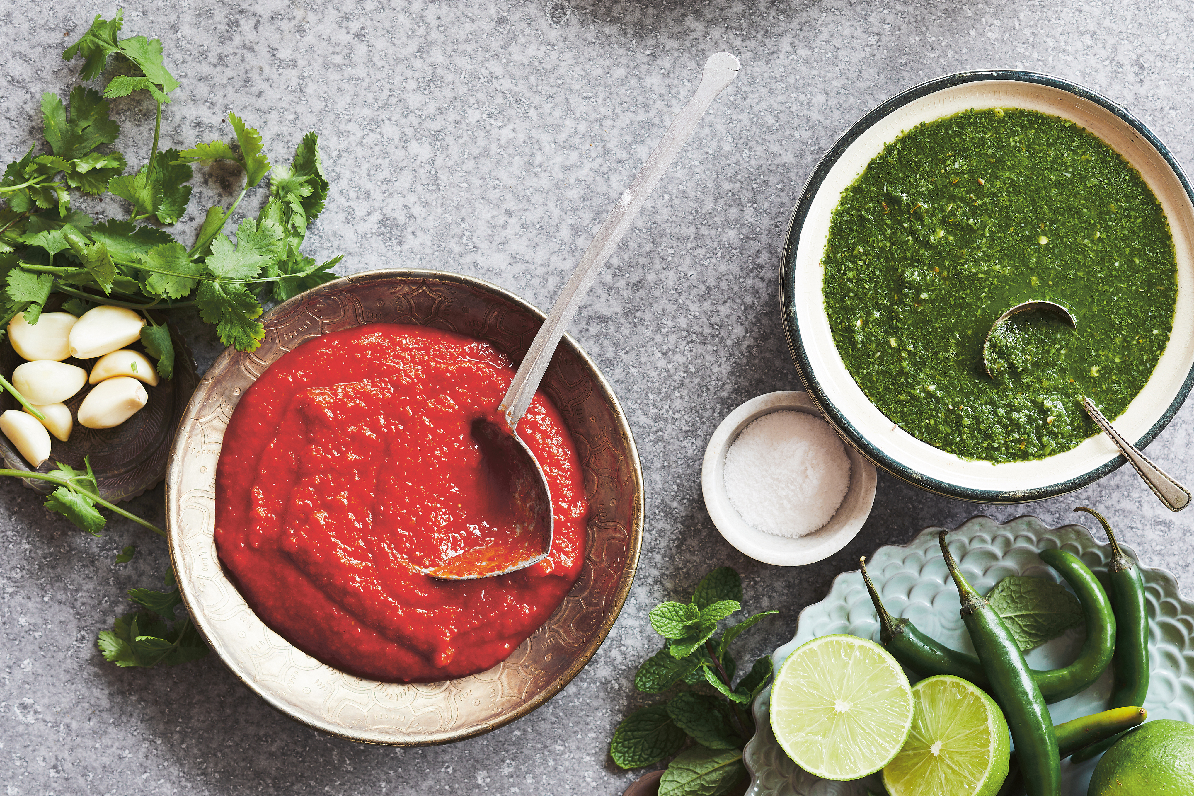 Overhead photo of a bowl of green chutney, a bowl of red chutney, and varied garnishes