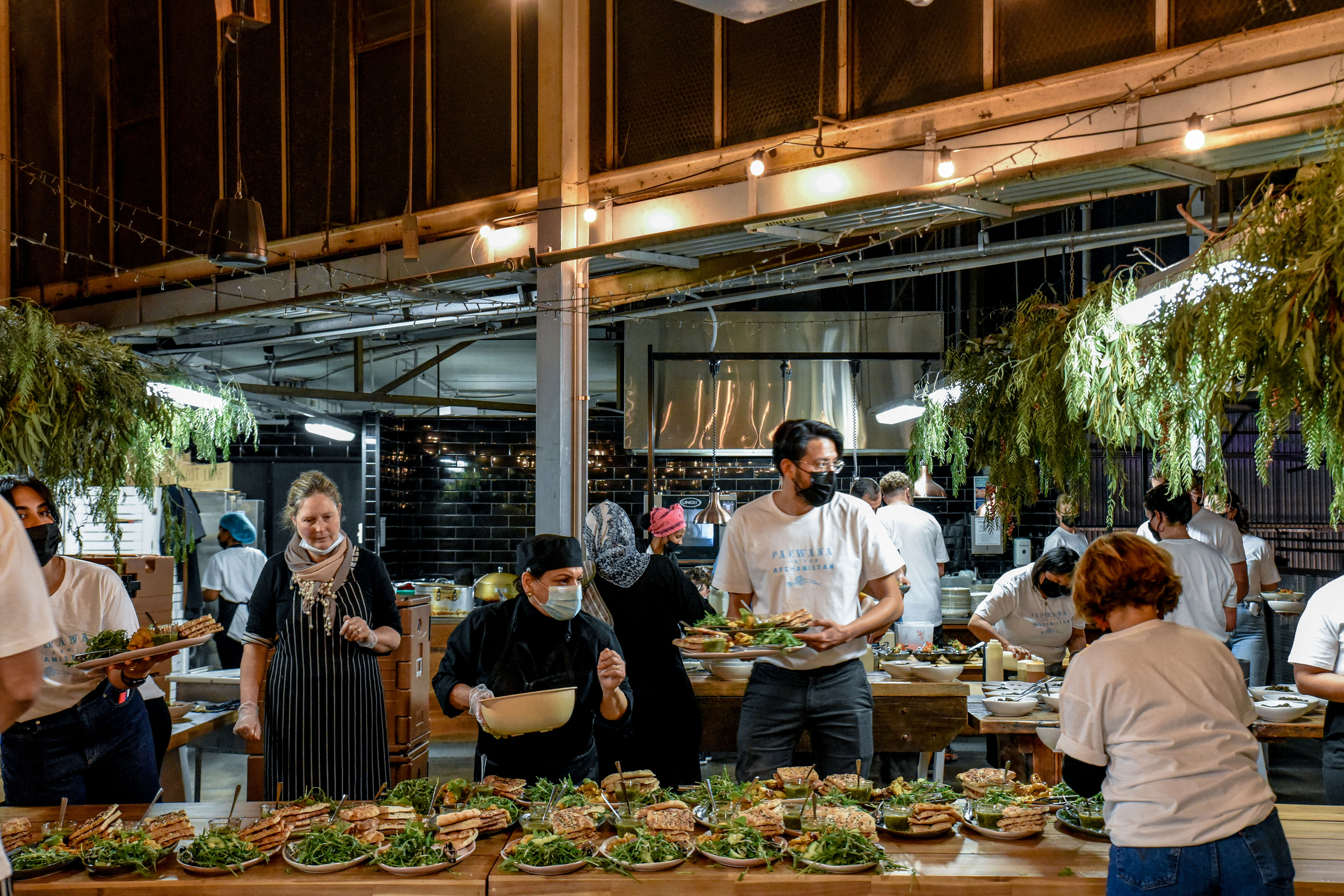 A long table with peole working on both sides. The table has tens of platters of salad, naan, and kebab laid out ready to go