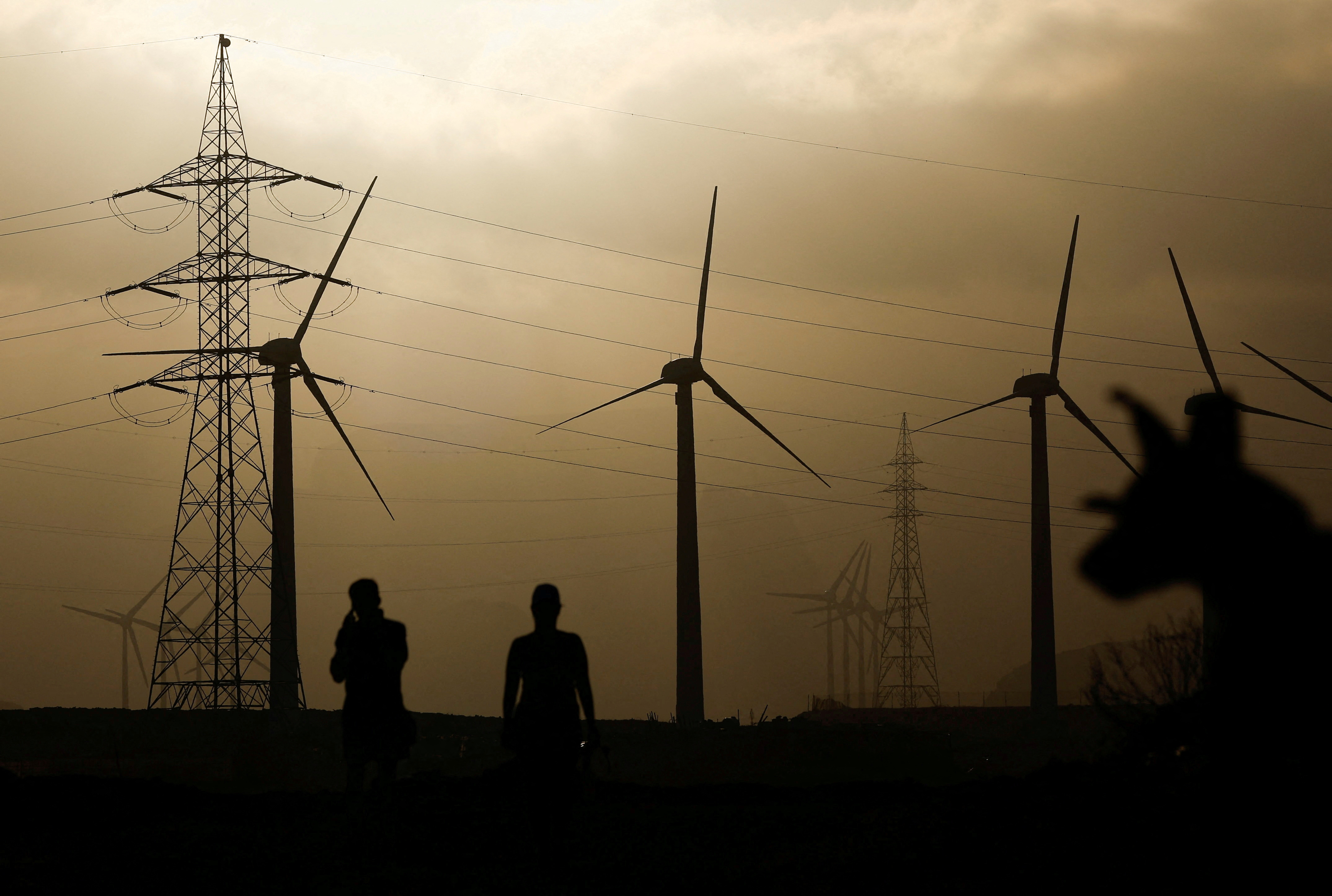 Two people walk near an electricity pylon and a group of wind turbines, in Barranco de Tirajana, in the island of Gran Canaria, Spain, May 11, 2022
