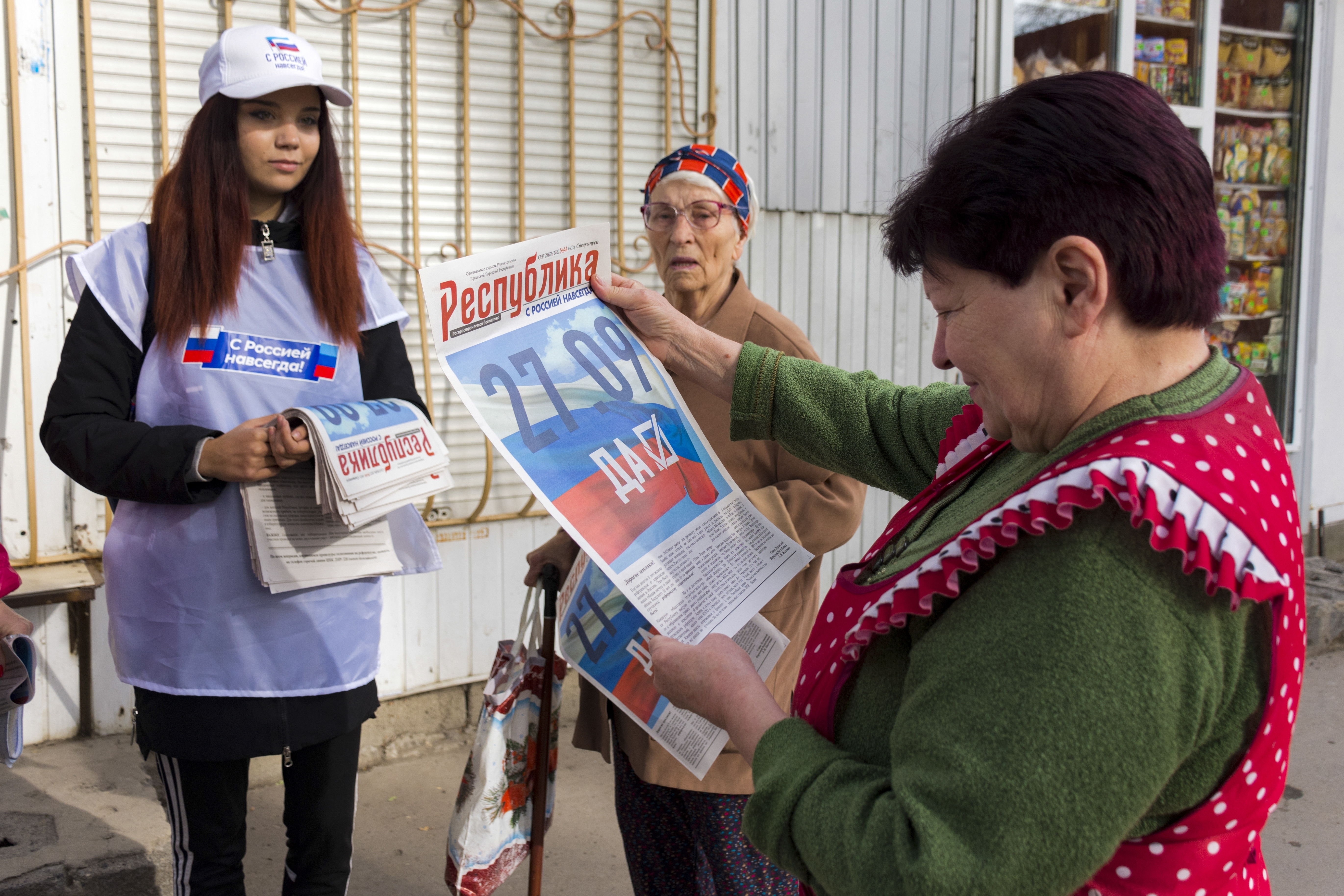 A woman looks at a newspaper ahead of the vote to join Russia that is due to start on Friday, watched by a young woman who is distributing the papers and an elderly woman
