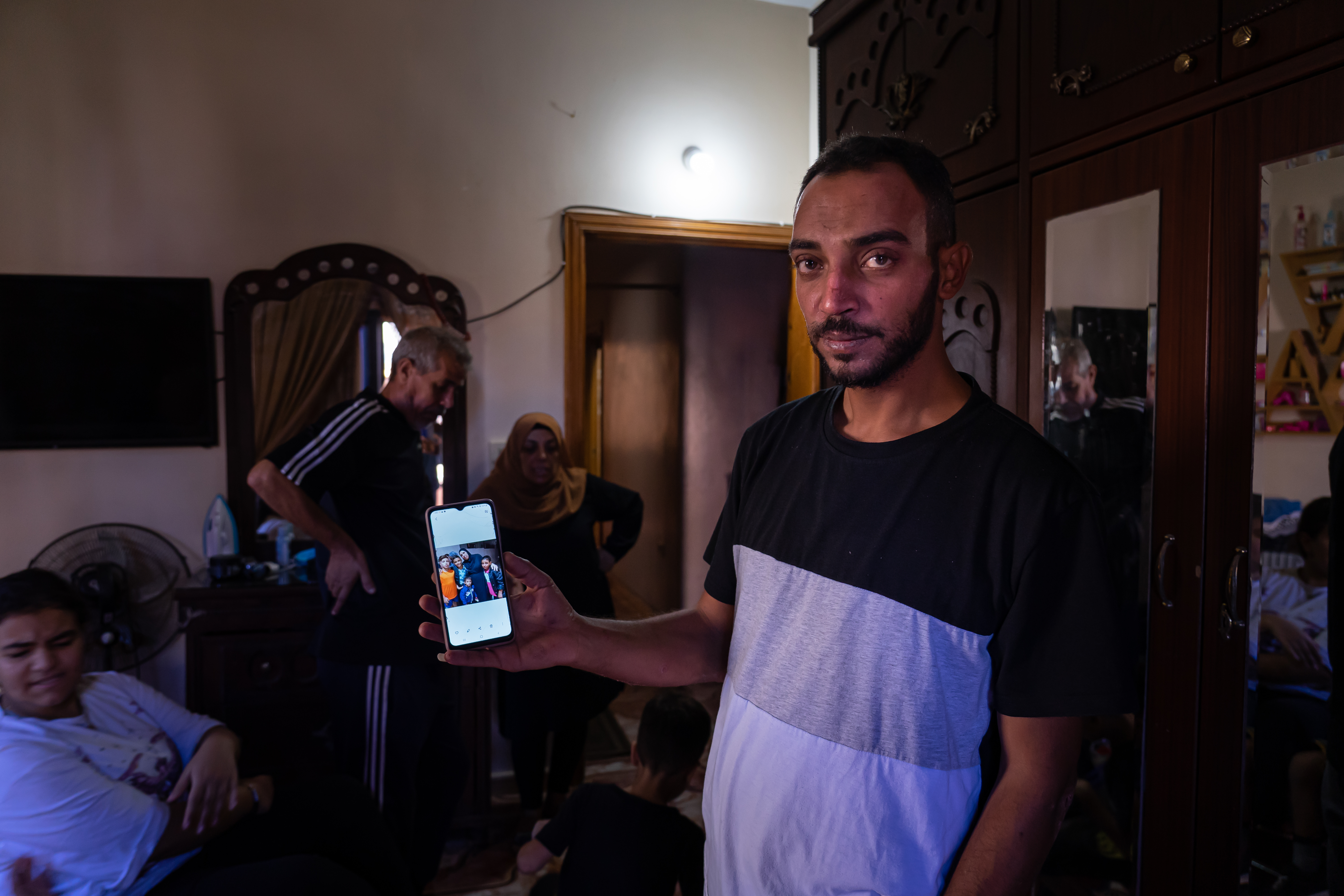 A man shows a picture on his phone in a dark room, others are in the background