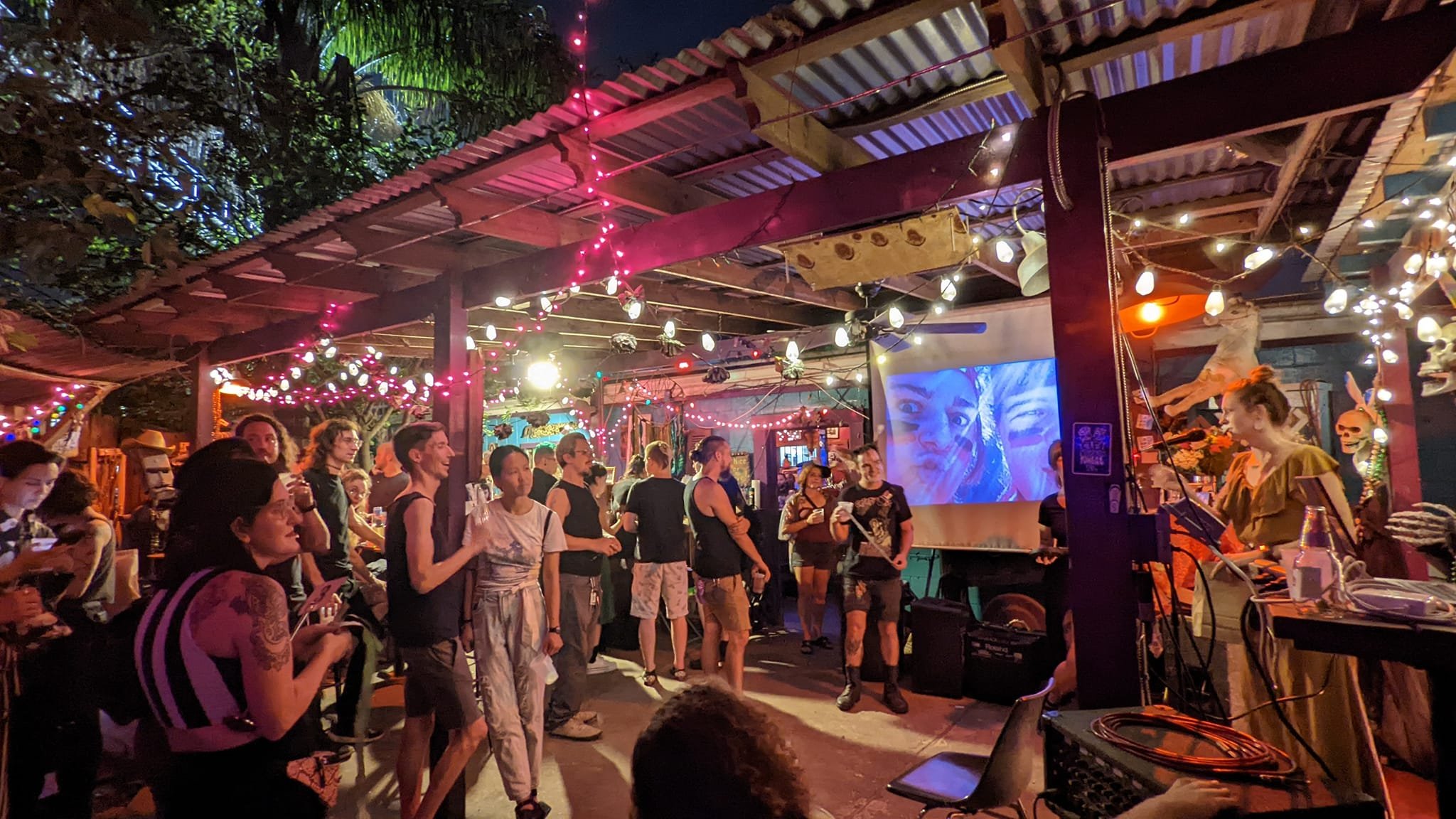 A photo of a crowd of people outdoors and a shelter with a metal roof with a photo or video being projected on a sheet in the shelter and the whole place is lit up using strings of light.