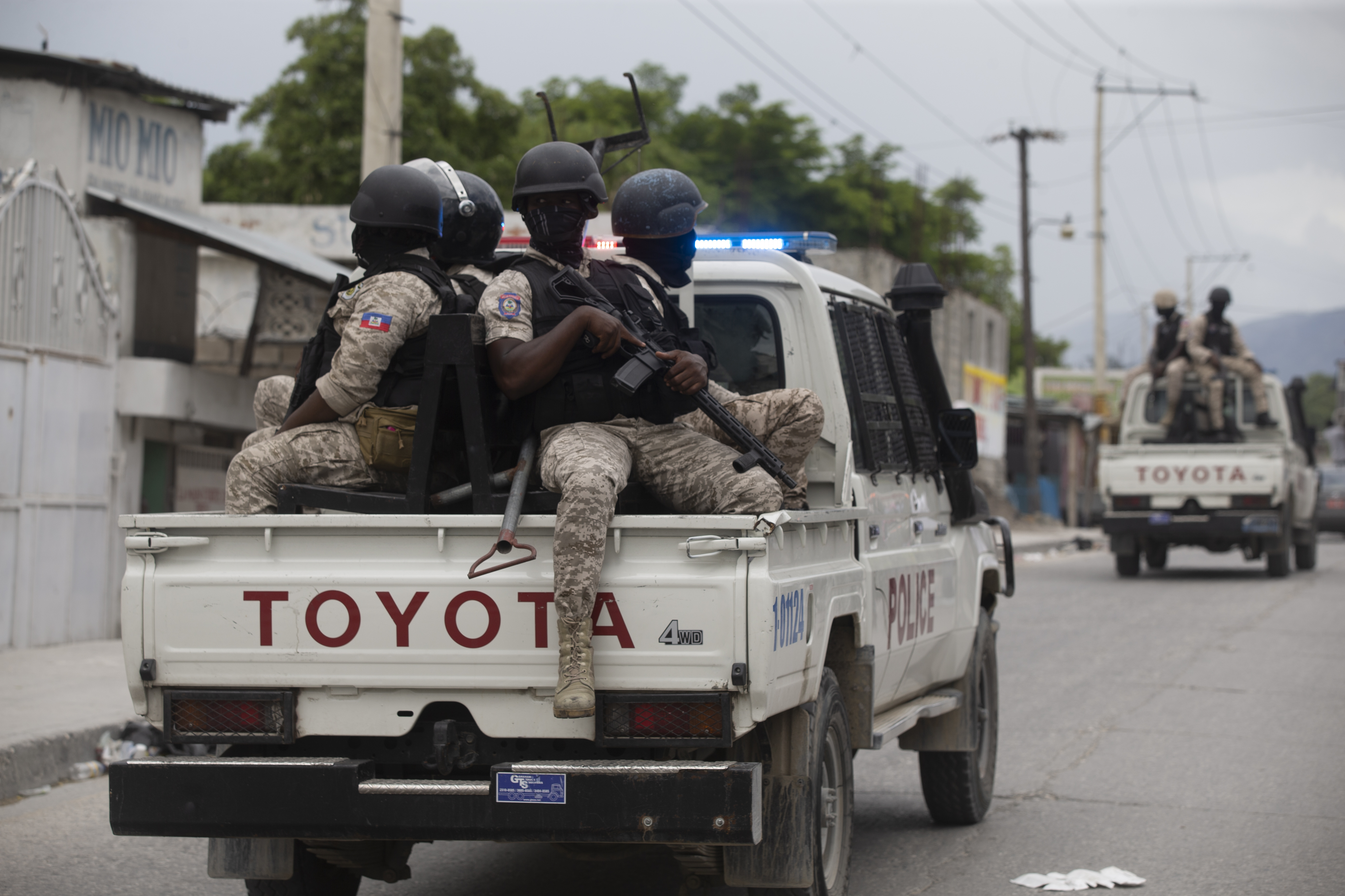 Security forces sit in the back of a white Toyota truck holding rifles and wearing black helmets and face gear. Another Truck is in the background in front of them, with forces also sitting in the back. The trucks say 'Police' on the side
