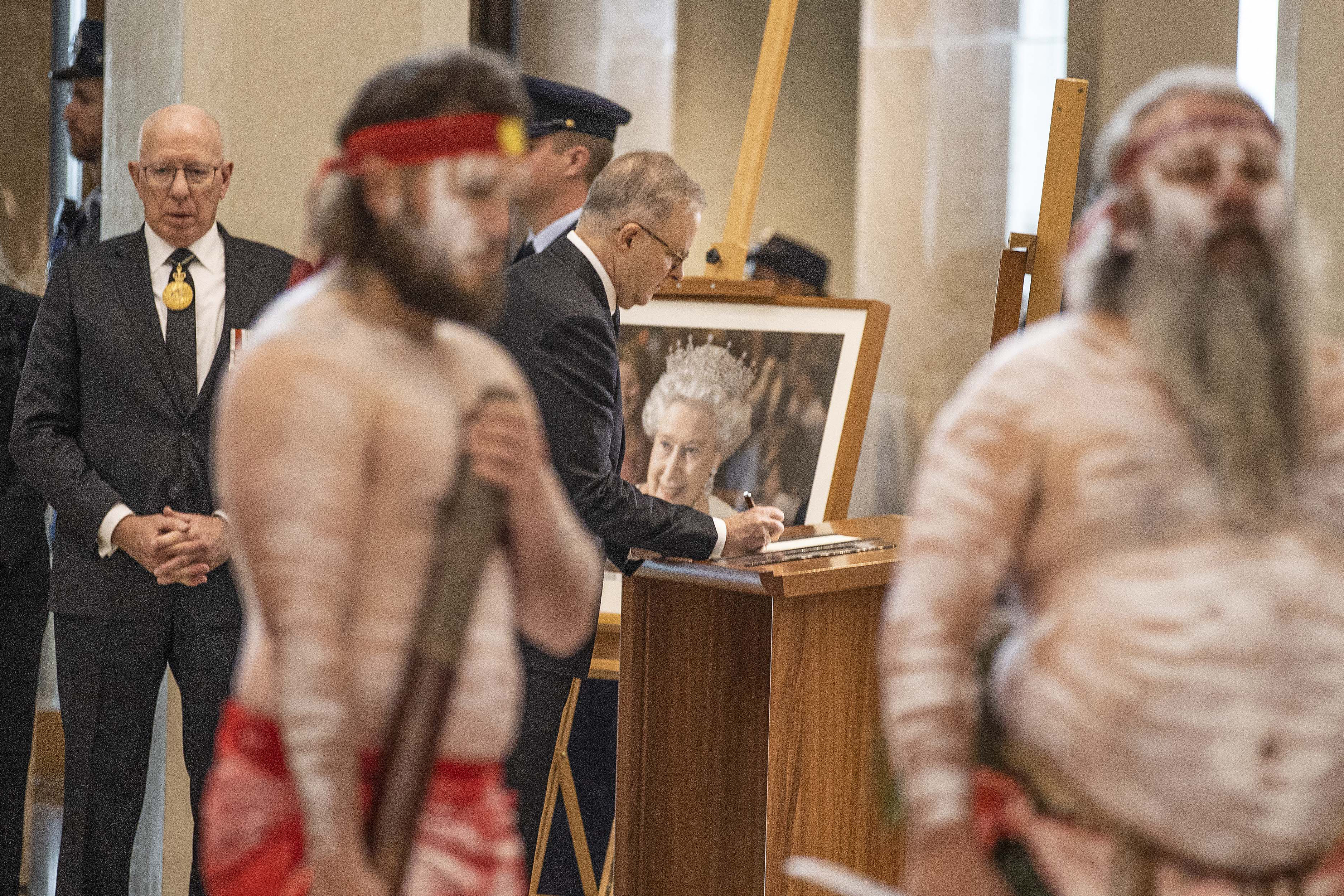 Auatralian PM Anthony Albanese signing a book in front of a picture of the queen with two Aboriginal men standing in the foreground